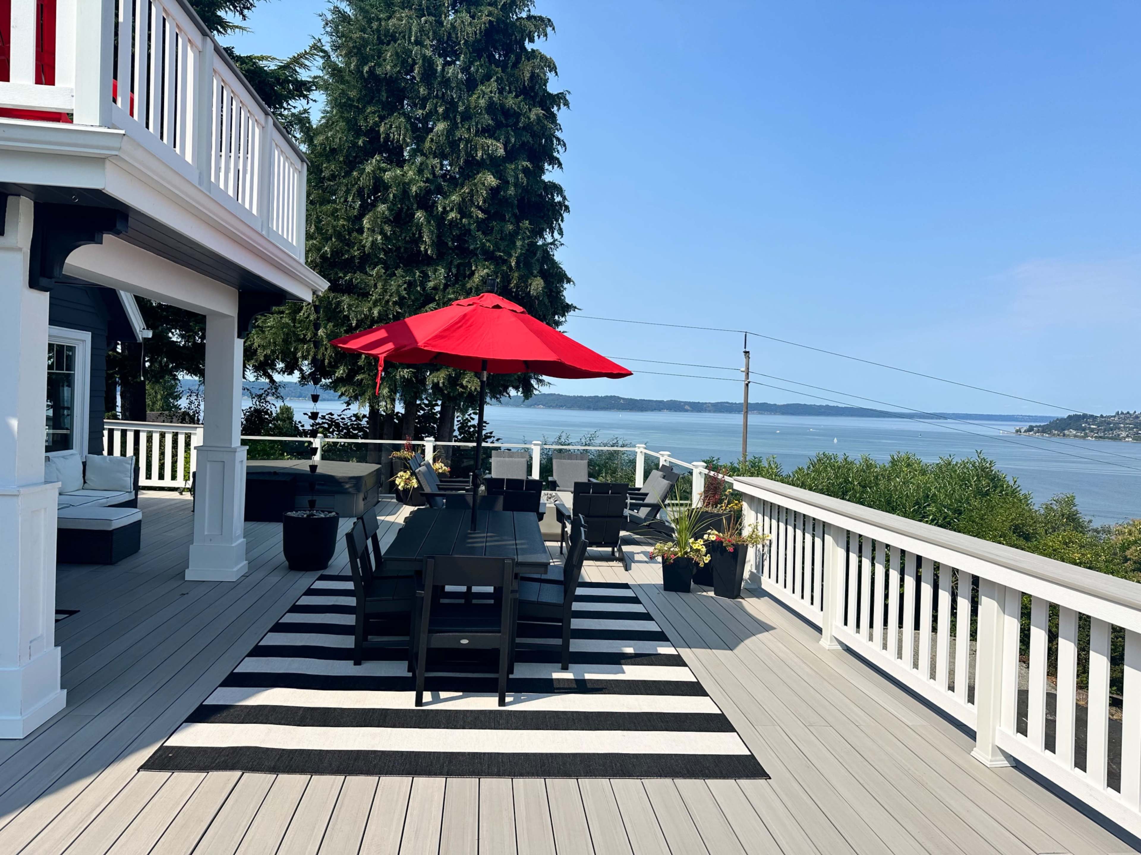 A spacious deck with black and white striped rugs features a dining area shaded by a red umbrella, overlooking a view of the water and distant shoreline.