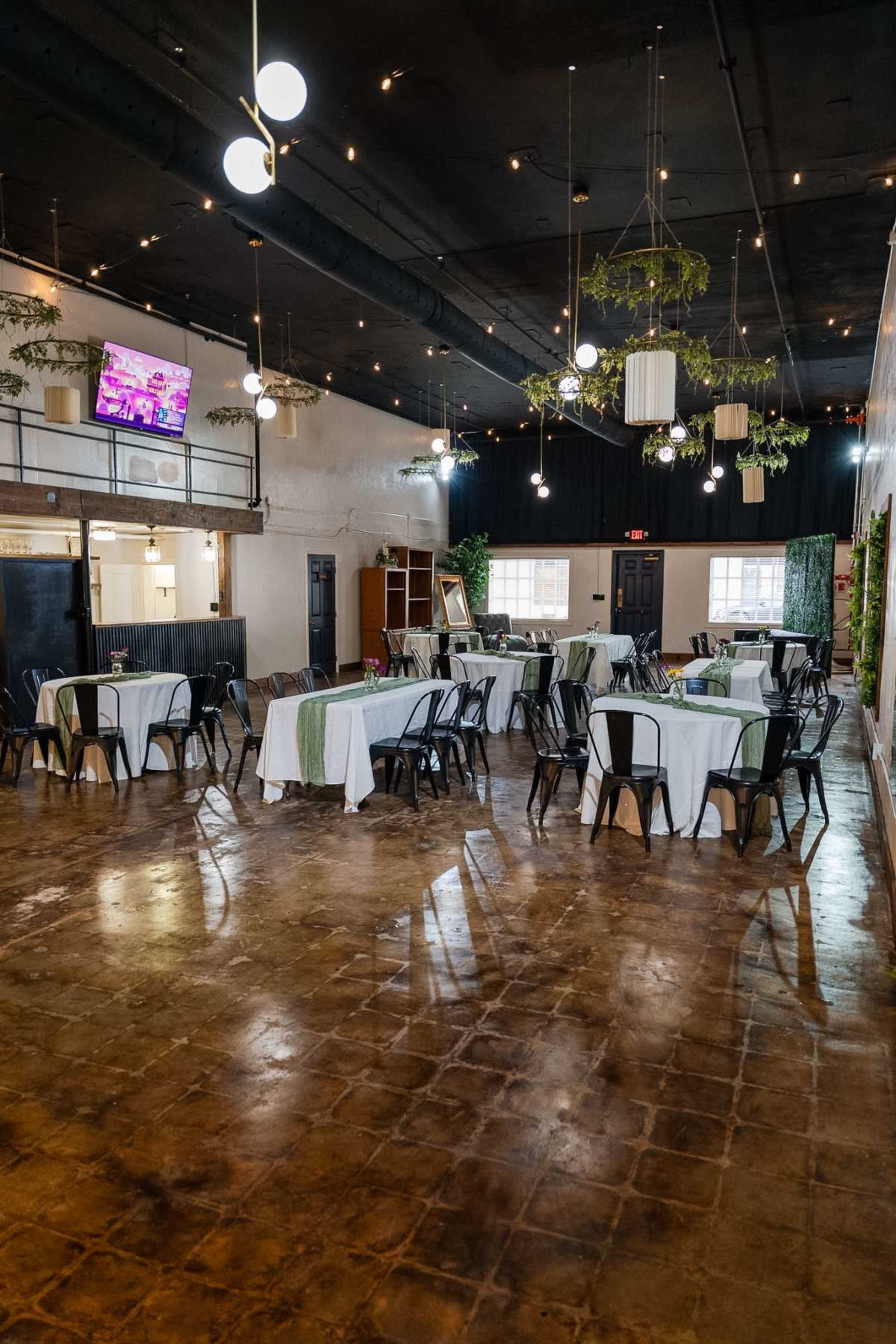 The image shows a spacious dining area with several tables covered in white tablecloths, black chairs, and modern lighting fixtures hanging from the ceiling.