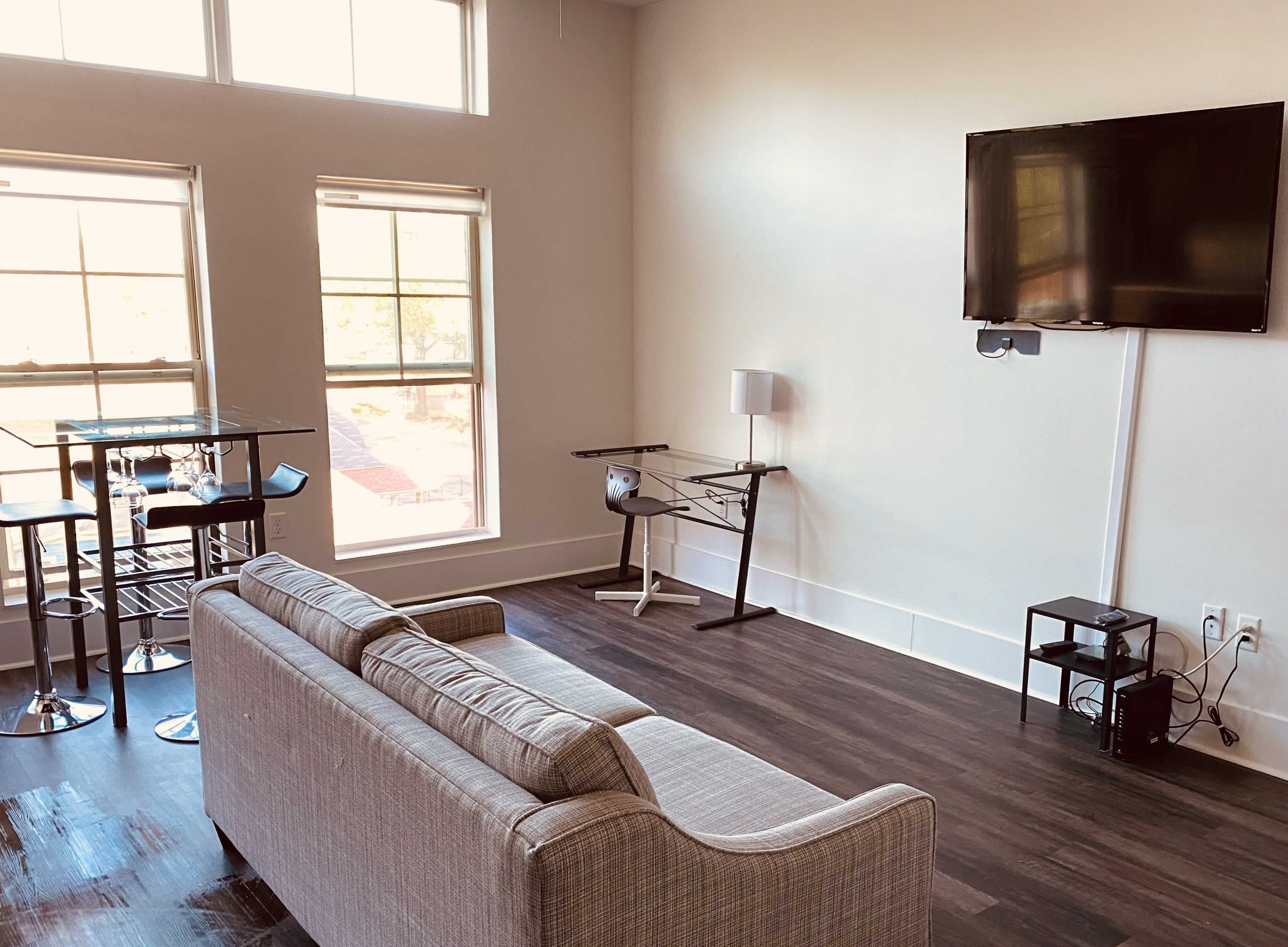 A spacious living area with a gray sofa, a glass dining table, a mounted television, and a small desk beside a window.