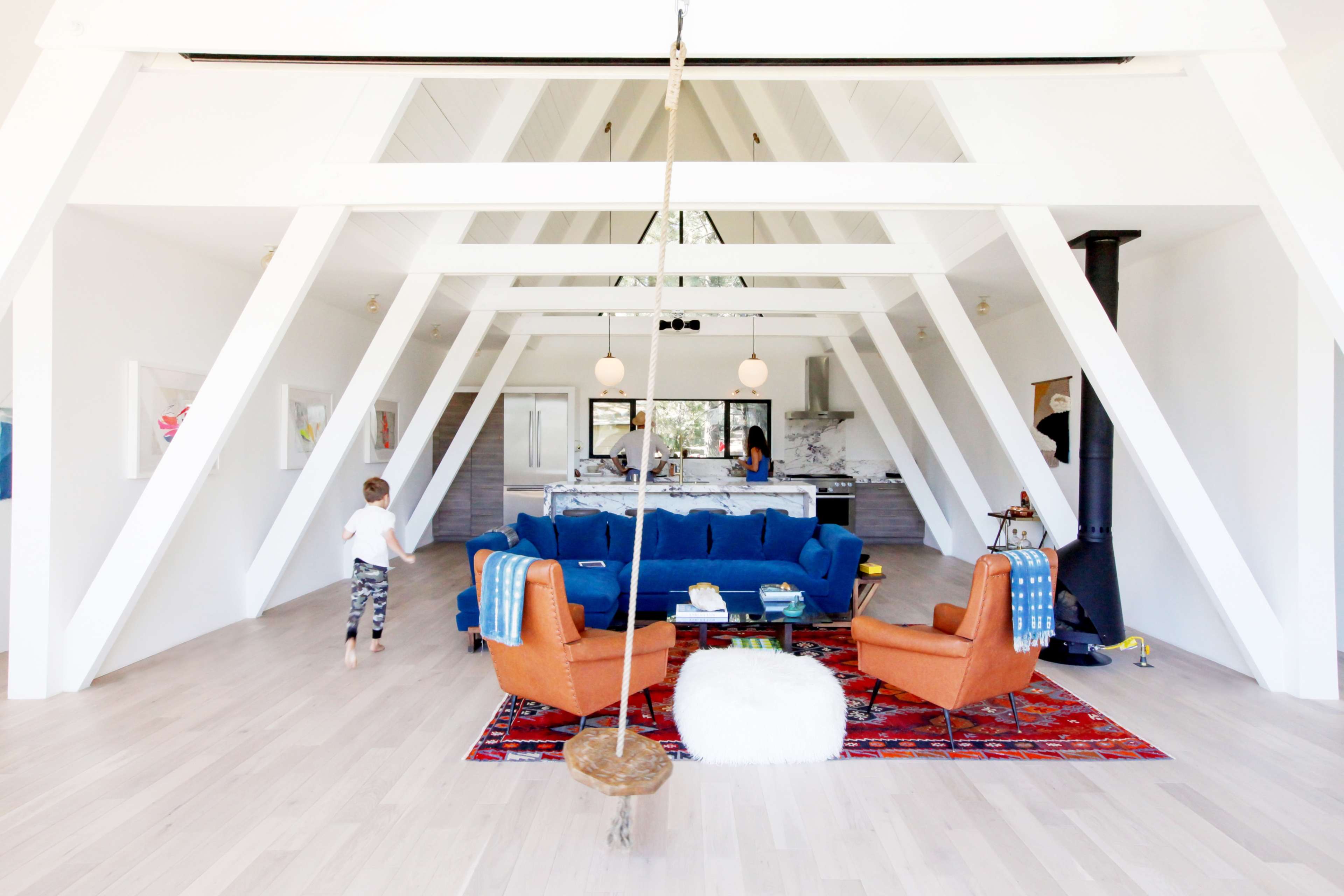 A spacious, modern living area with a blue sectional sofa, orange armchairs, a white rug, a swing, and a kitchen visible in the background.