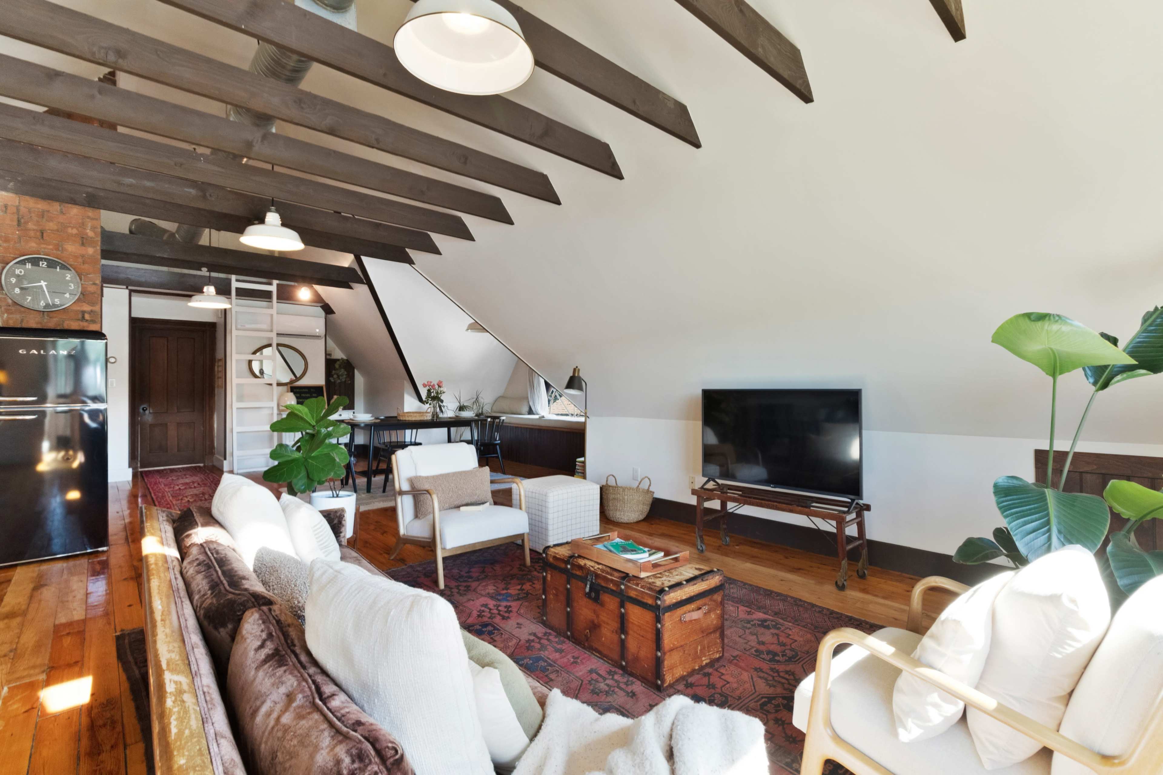 A cozy living room features wooden beams, a television, and a mix of furniture including a sofa, two armchairs, and a coffee table in front of a carpet.