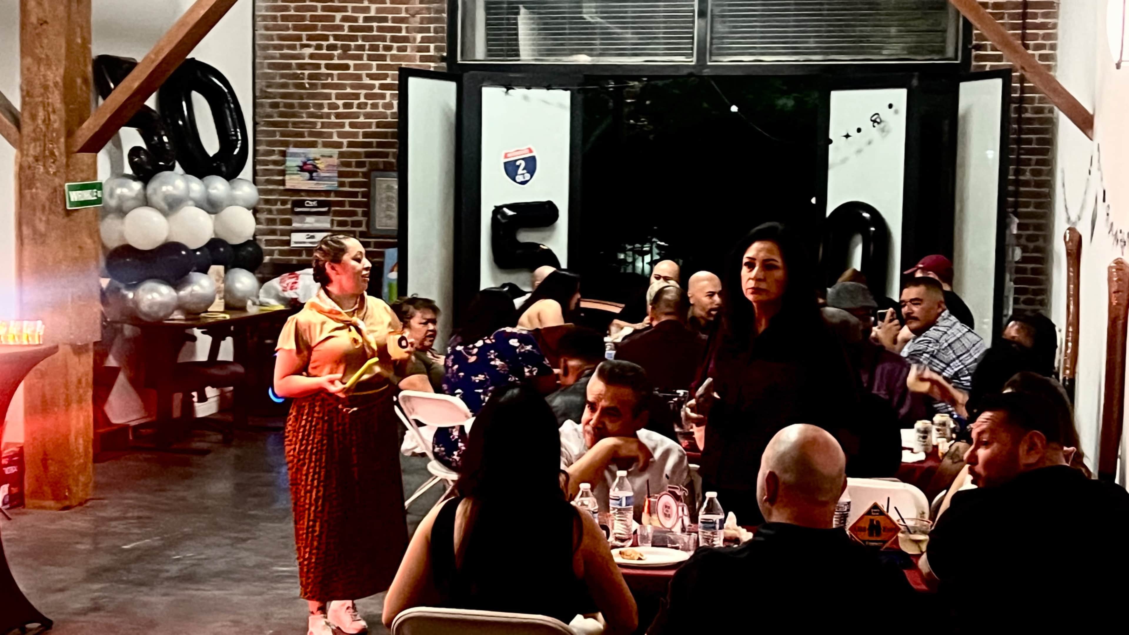 A group of people is seated at tables during a gathering in an industrial-style venue with exposed brick walls and balloons in the background.