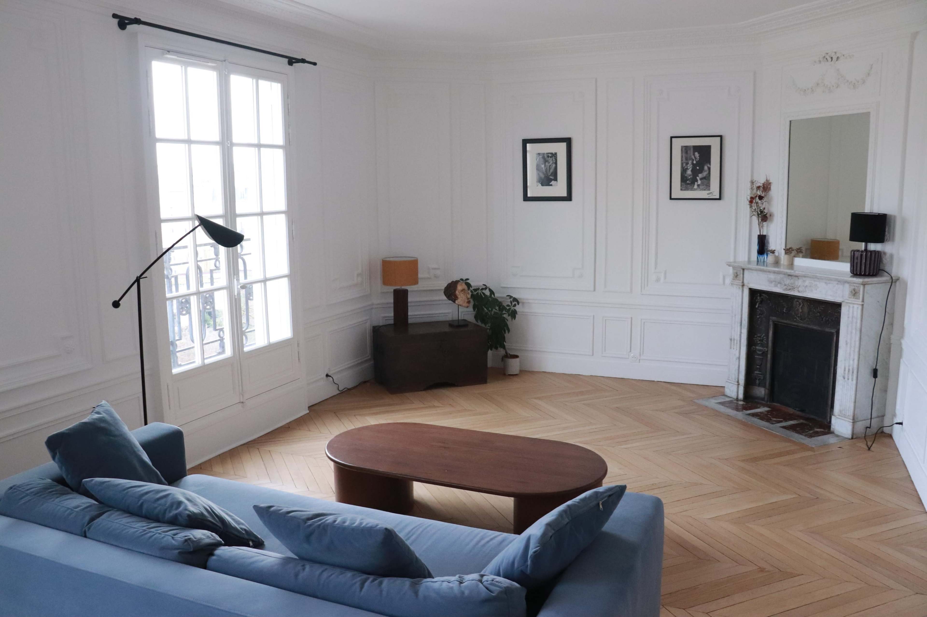 The image shows a bright, empty living room with a blue sofa, a wooden coffee table, a lamp, and decorative elements along the white walls and hardwood floor.