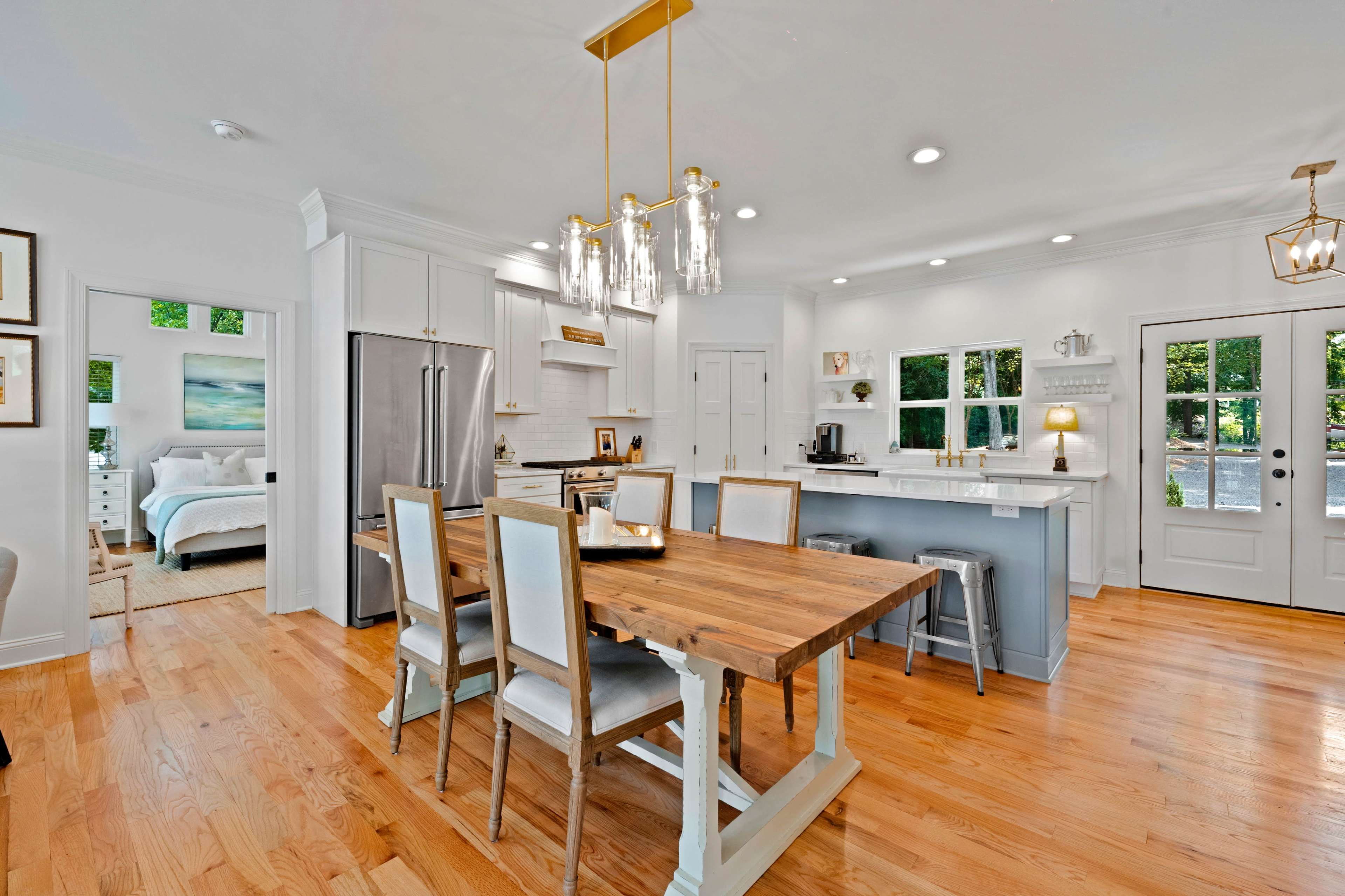 A modern kitchen and dining area feature a wooden table, white cabinetry, and stainless steel appliances, with a view of a well-lit adjoining room.
