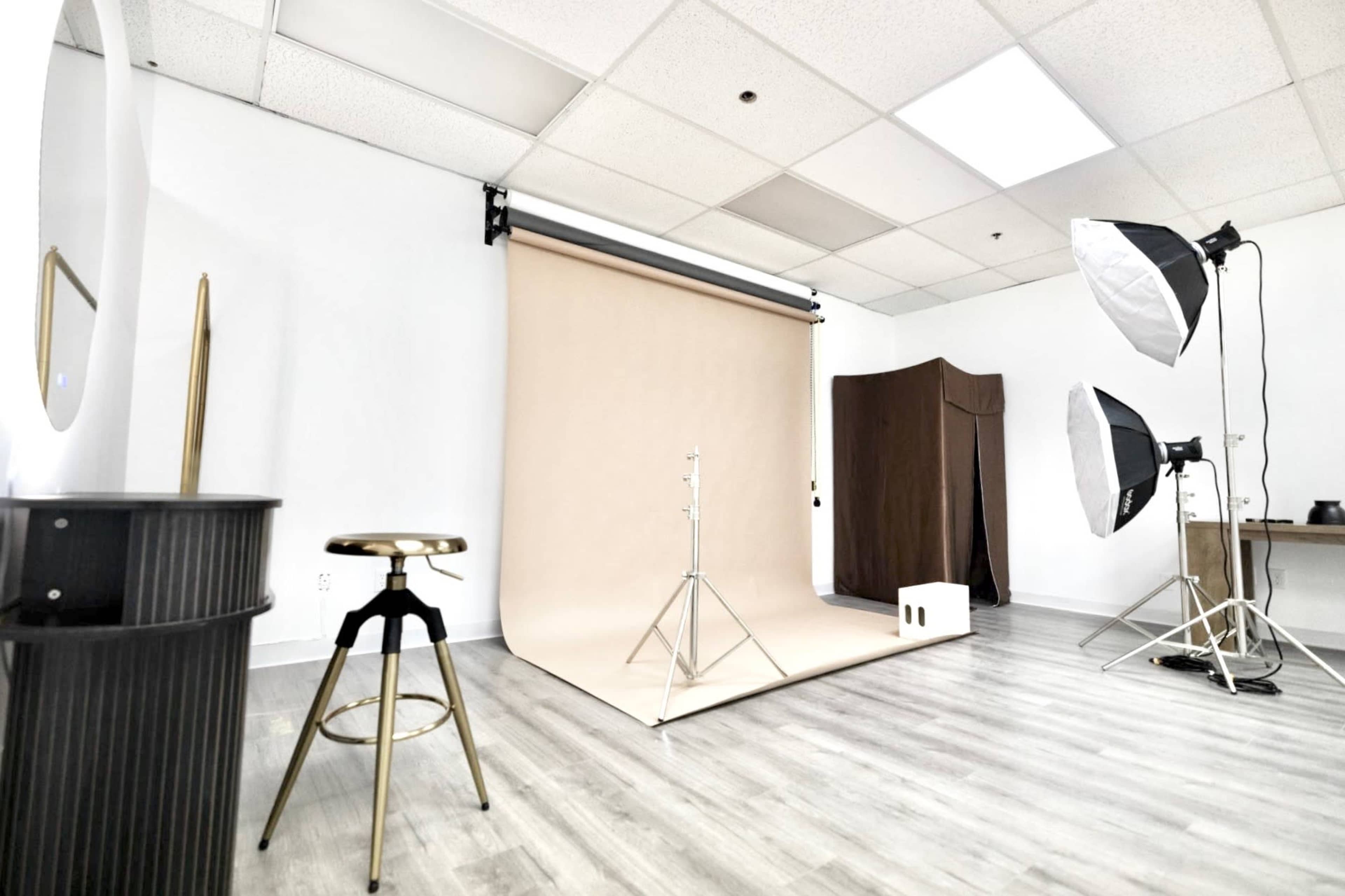 The image shows a photography studio featuring a beige backdrop, softbox lights, and a stool against a neutral wall.