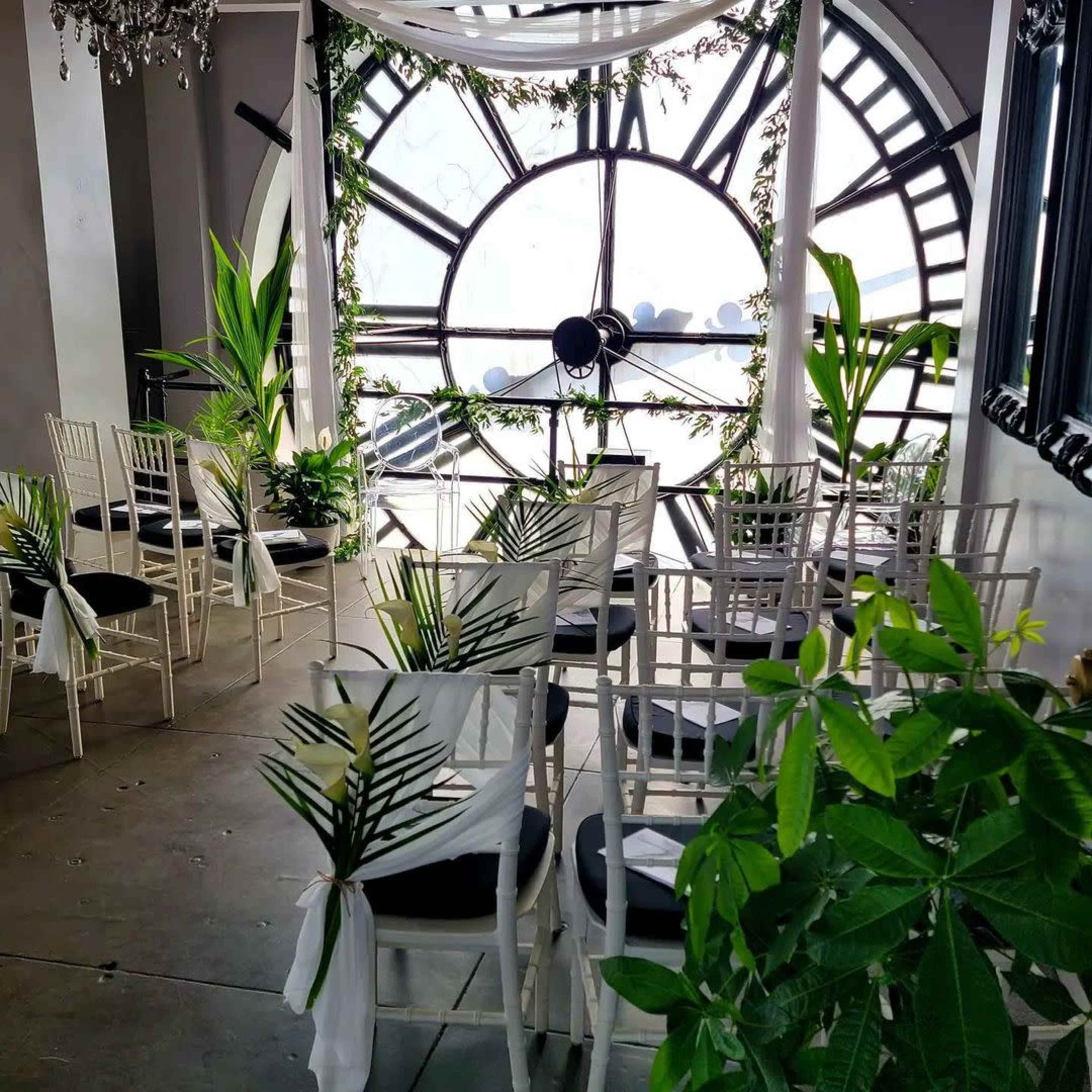A wedding ceremony setup with white chairs adorned with greenery, arranged in front of a large clock window.