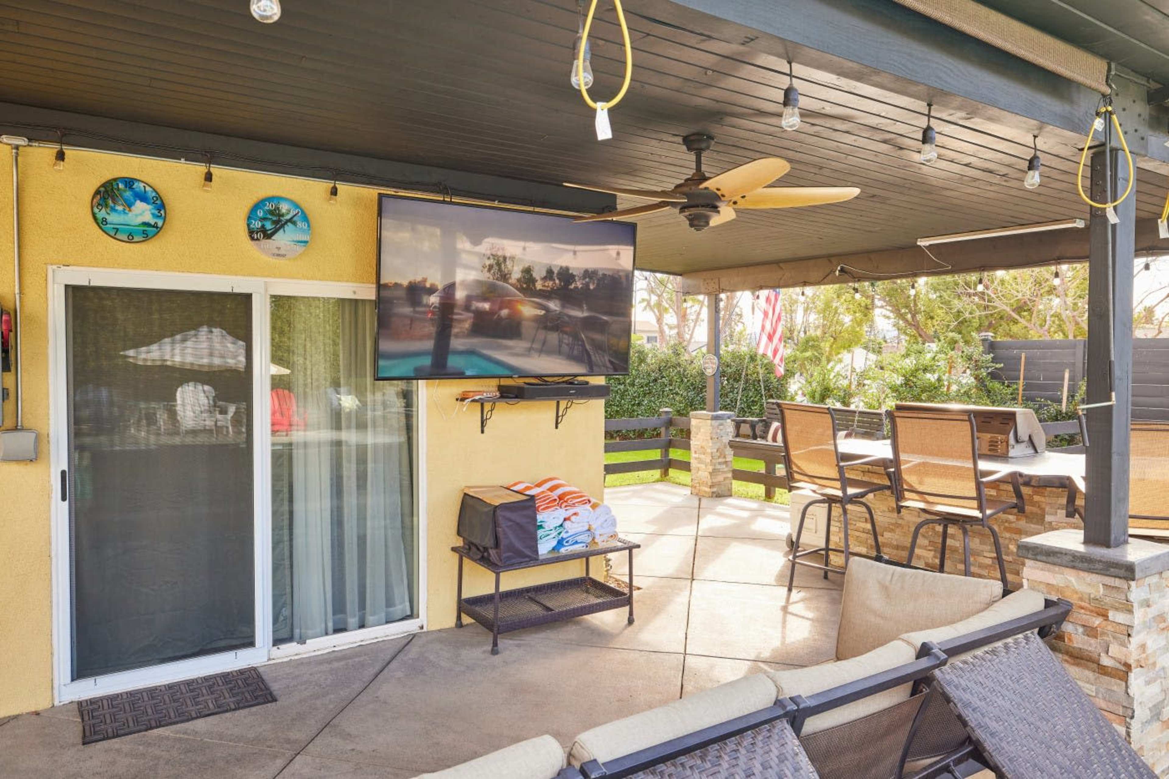 The image shows a patio area with a mounted TV, ceiling fan, and outdoor seating, surrounded by greenery and notable decor.