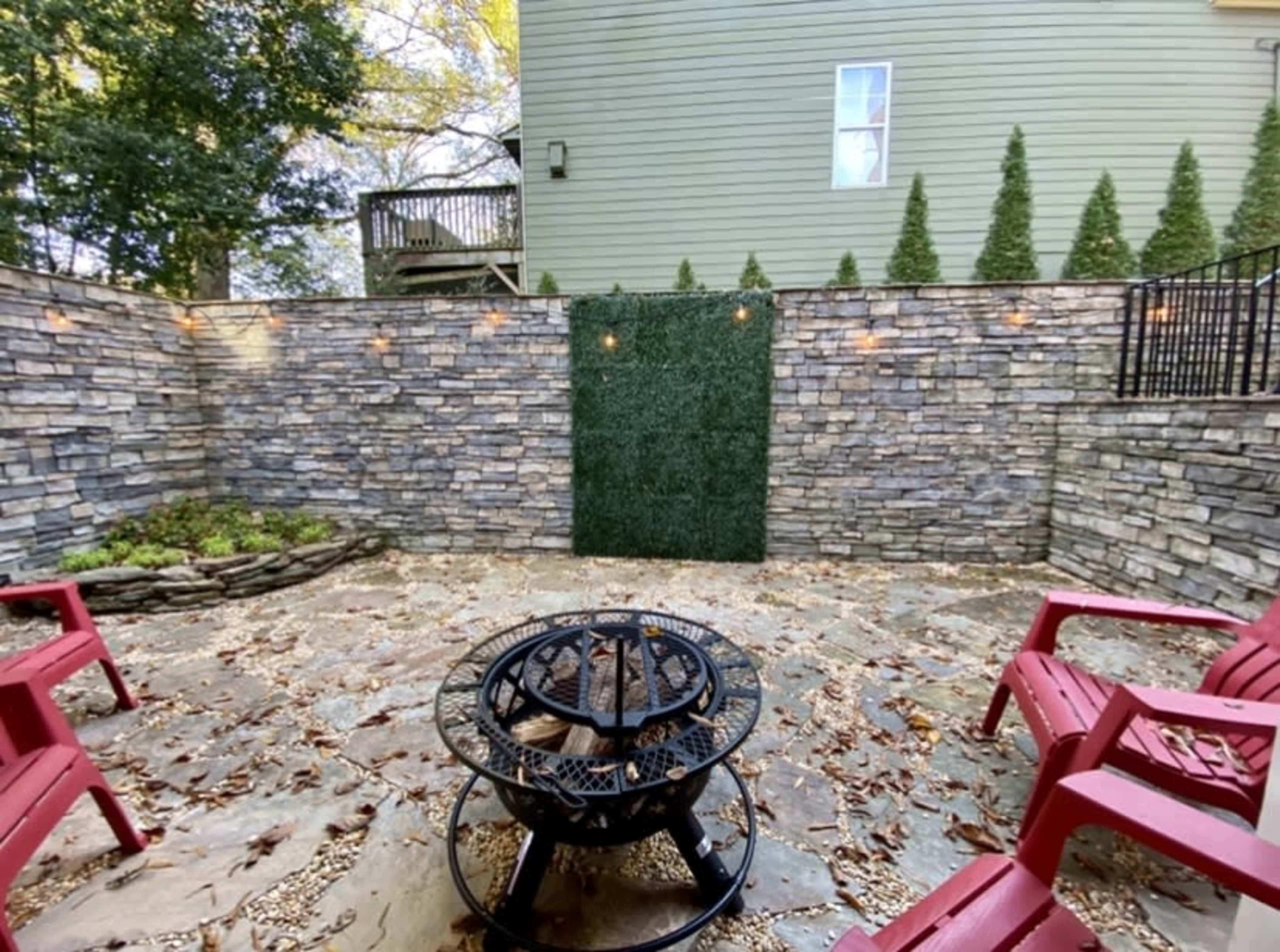 A backyard area with a stone-paved surface, surrounded by a tall stone wall and featuring a circular fire pit at the center, with red chairs arranged around it.