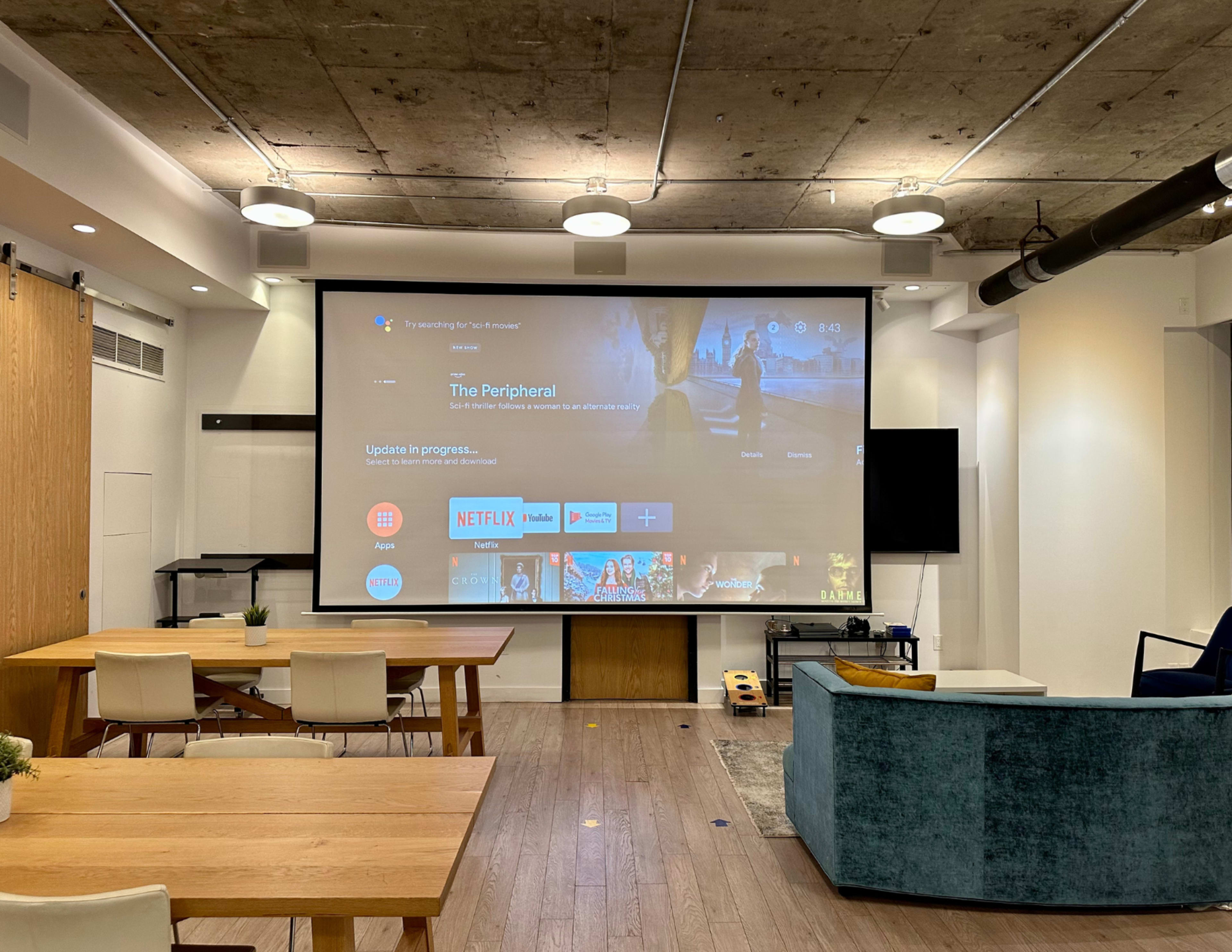 A large screen displaying a streaming service interface is set up in a modern room with wooden tables and a couch.