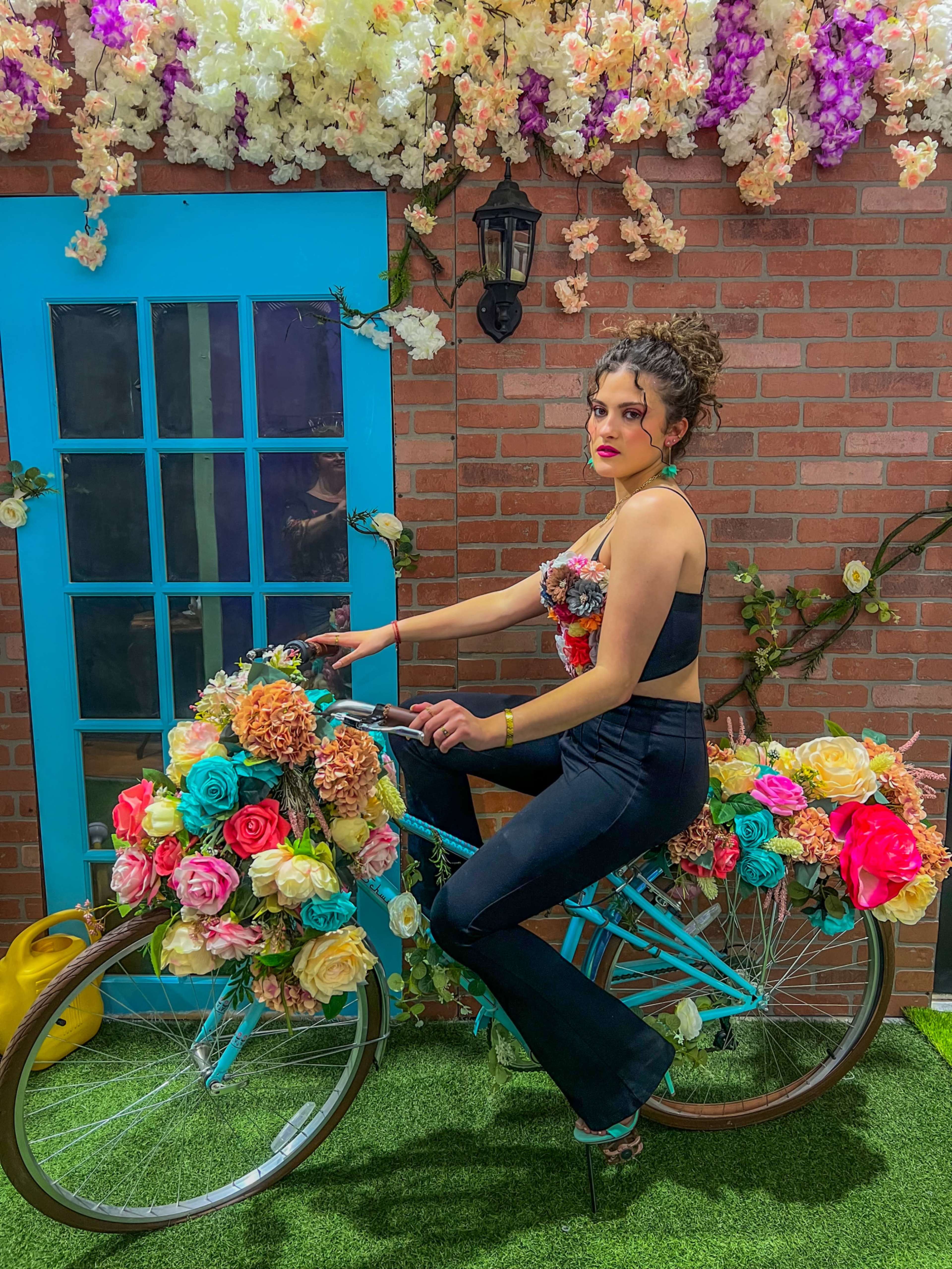 A woman poses on a flower-decorated bicycle in front of a blue door and a brick wall surrounded by artificial plants.
