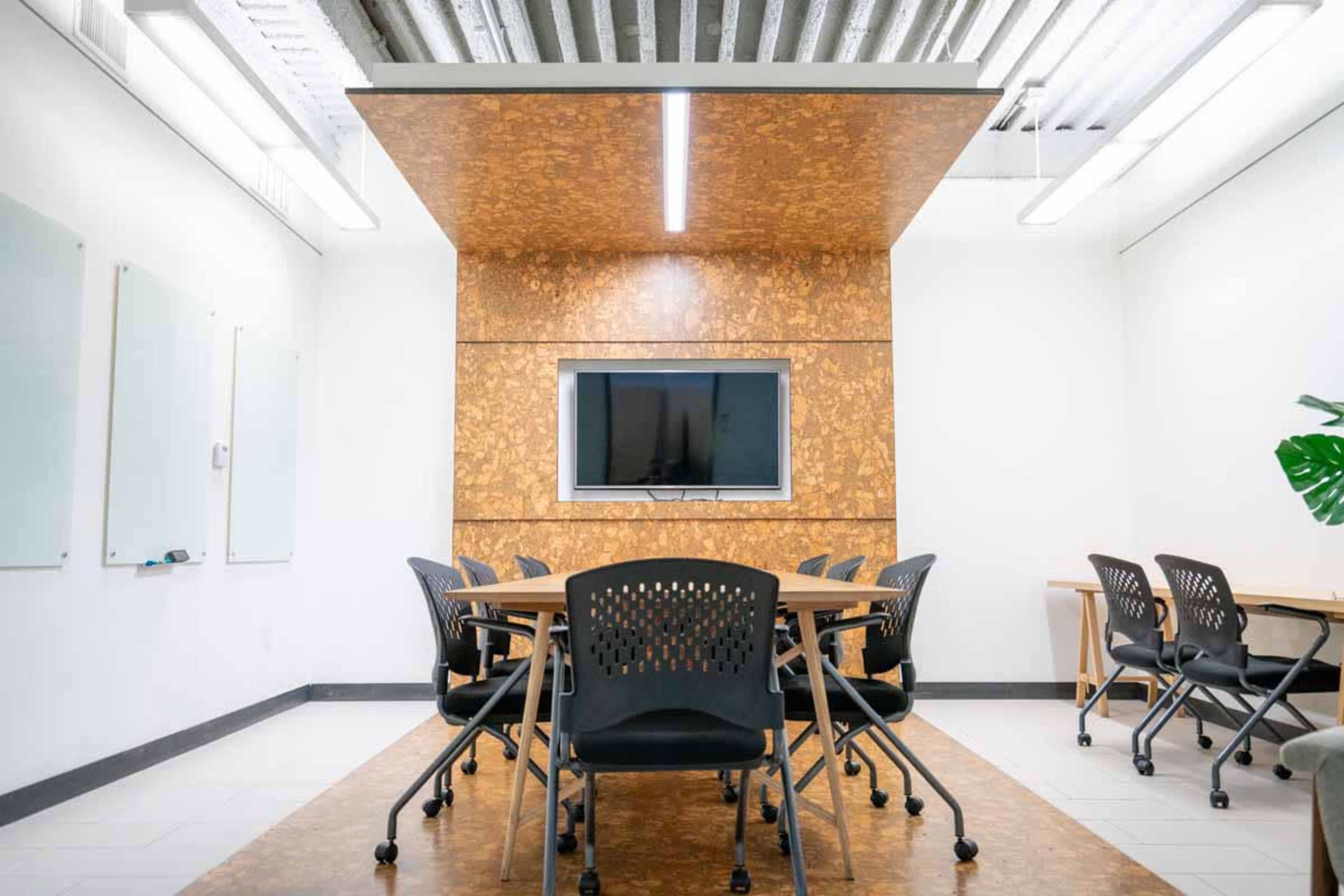 A modern conference room features a central table surrounded by black chairs, with a wall-mounted television and cork paneling in the background.