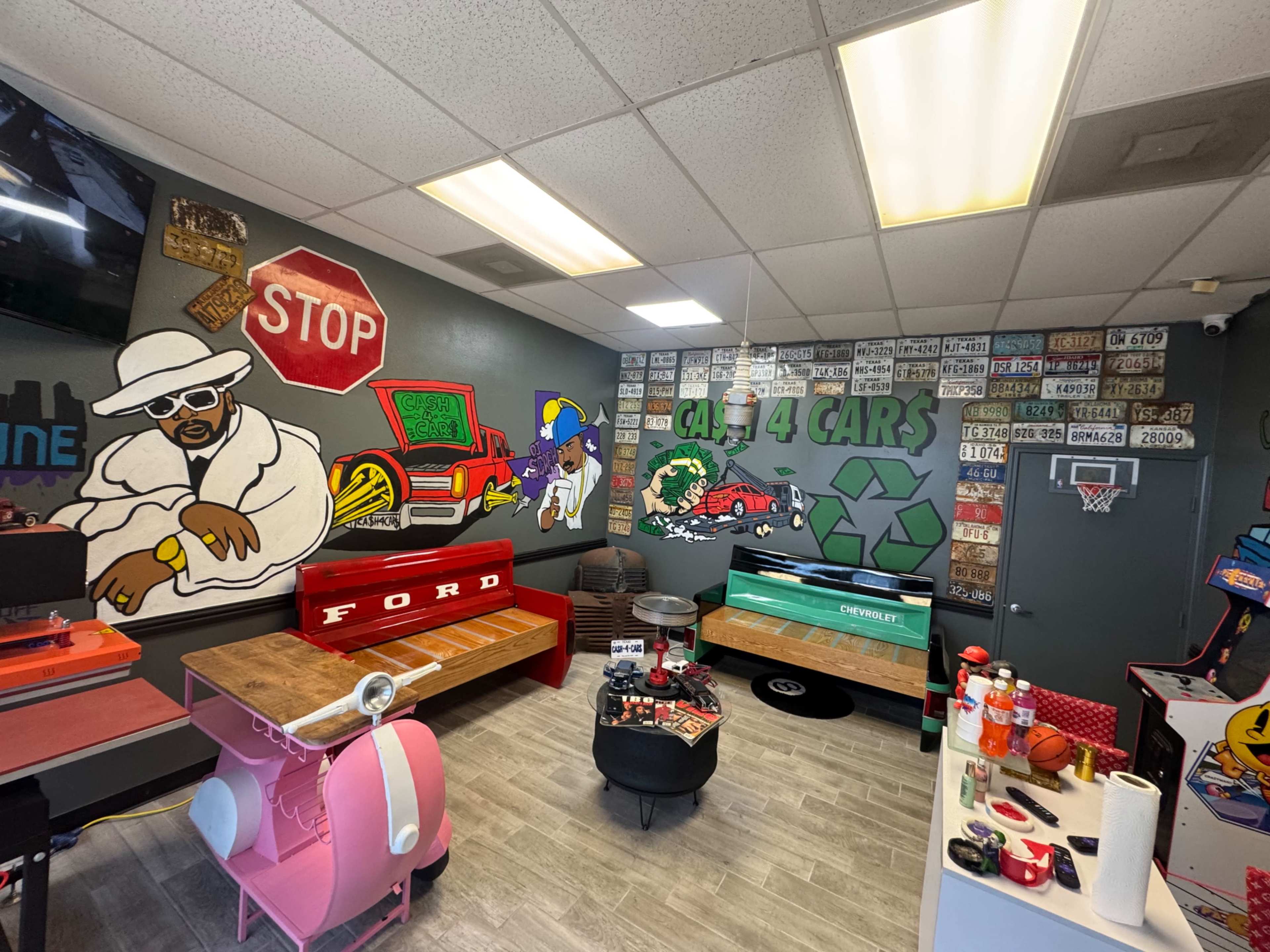 The image shows a vibrant auto-themed room featuring colorful murals, multiple vintage car-themed tables, and various license plates displayed on the walls.