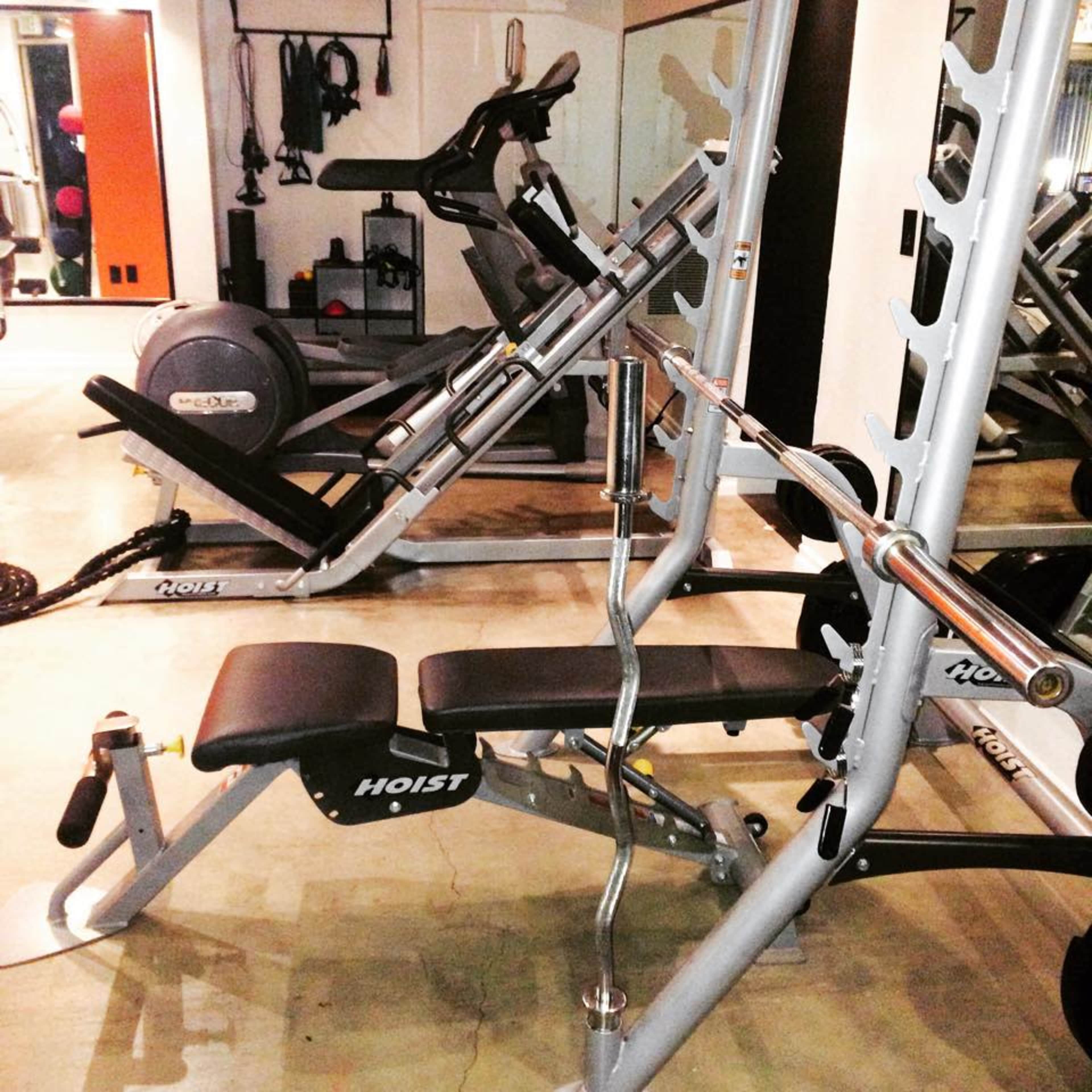 The image shows a gym interior with various workout equipment, including a bench press, exercise bike, and weight plates.