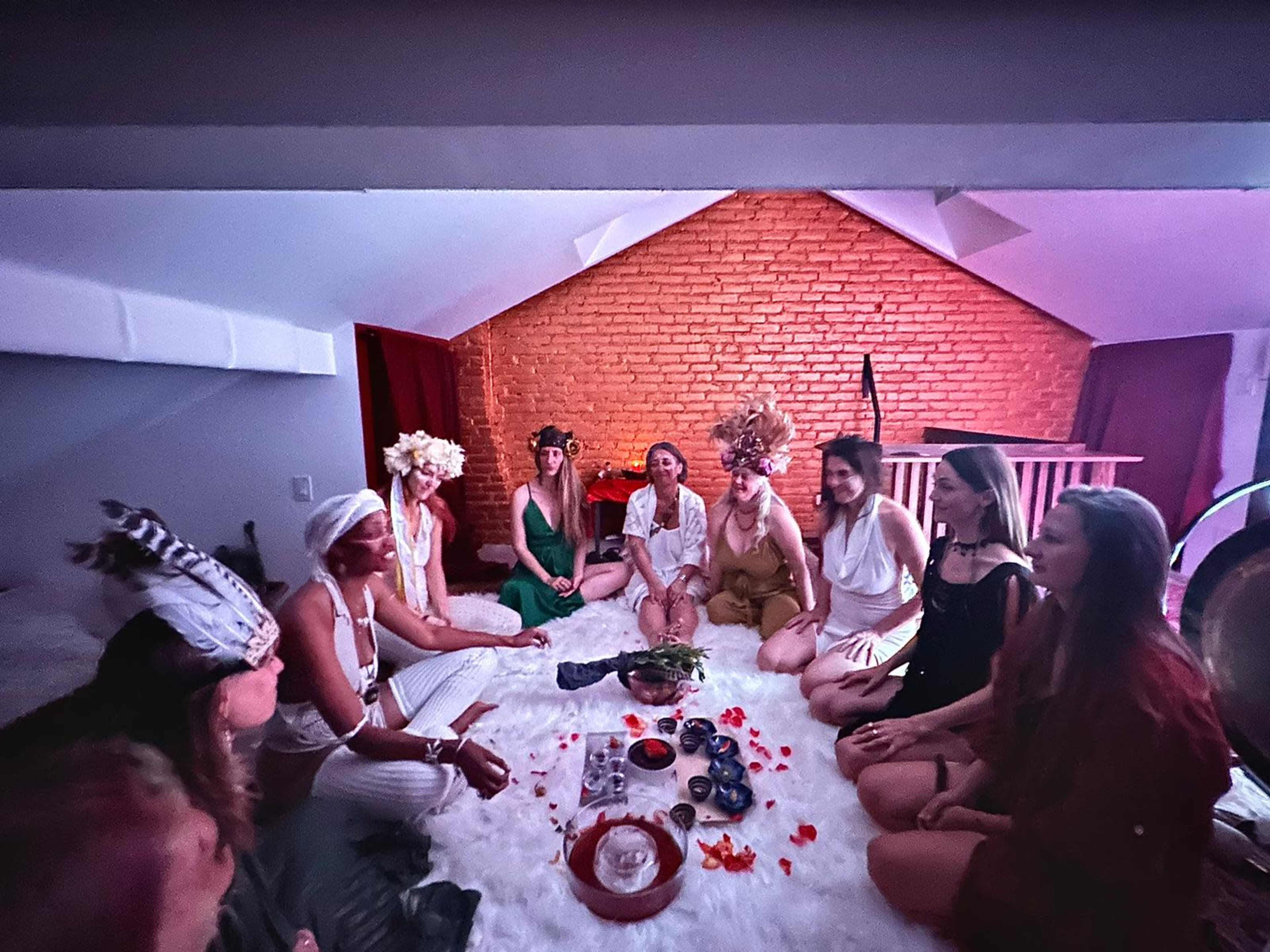 A group of women sits in a circle on a white rug in a room with a brick wall, engaged in a communal gathering with flowers and offerings in the center.