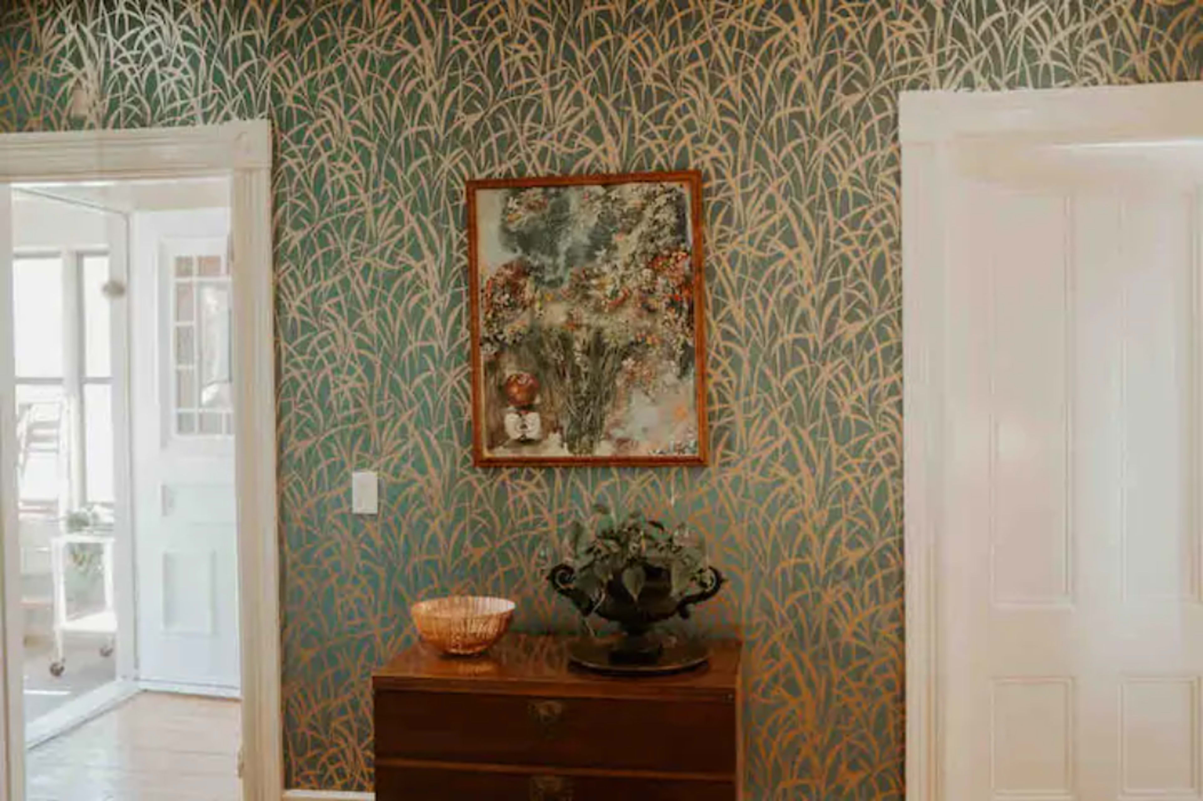 A room with patterned wallpaper featuring tall grasses, a framed artwork on the wall, and a wooden dresser topped with a decorative bowl and a potted plant.