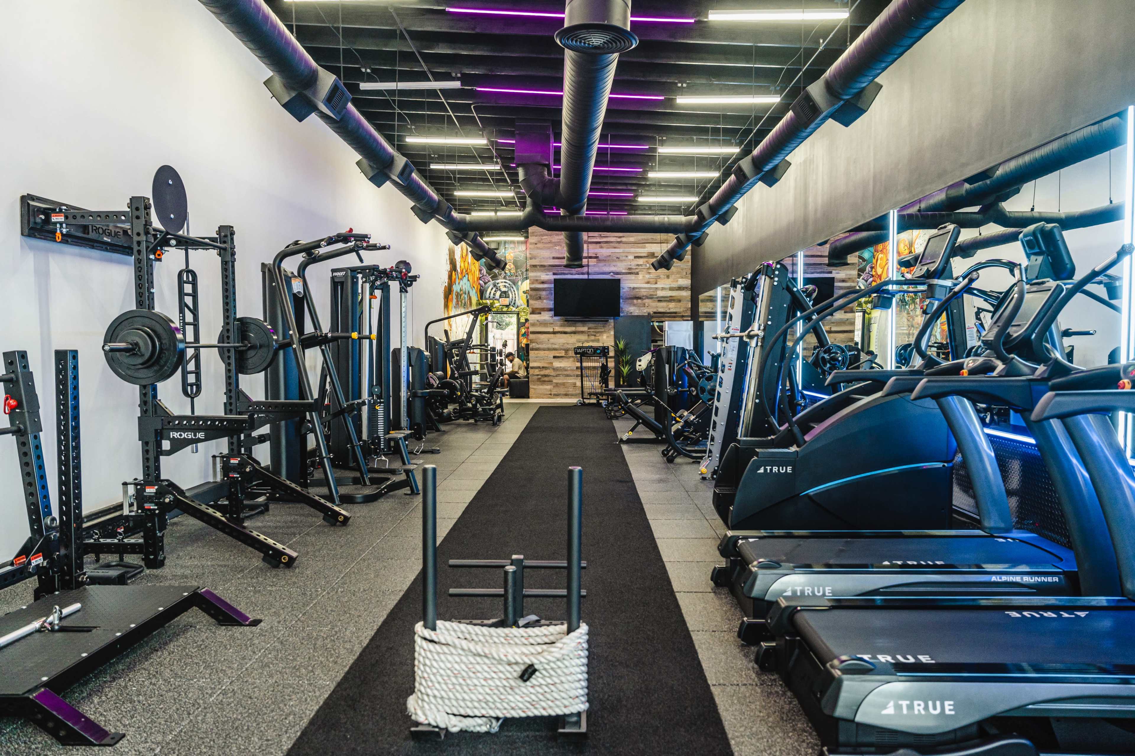 The image shows a modern gym interior equipped with various fitness machines, free weights, and exercise equipment arranged along a central walkway.
