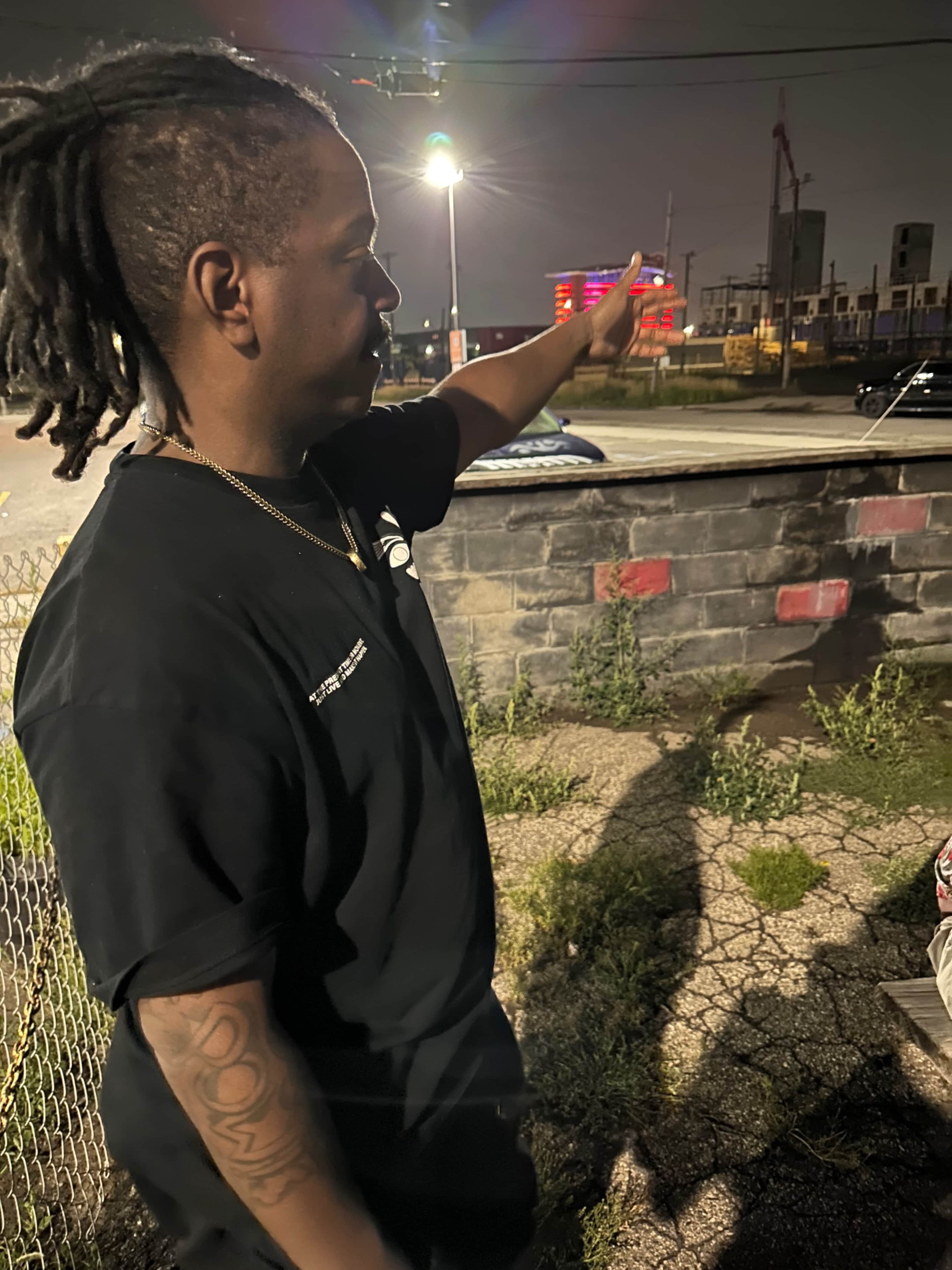 A man with dreadlocks gestures toward a construction site at night.