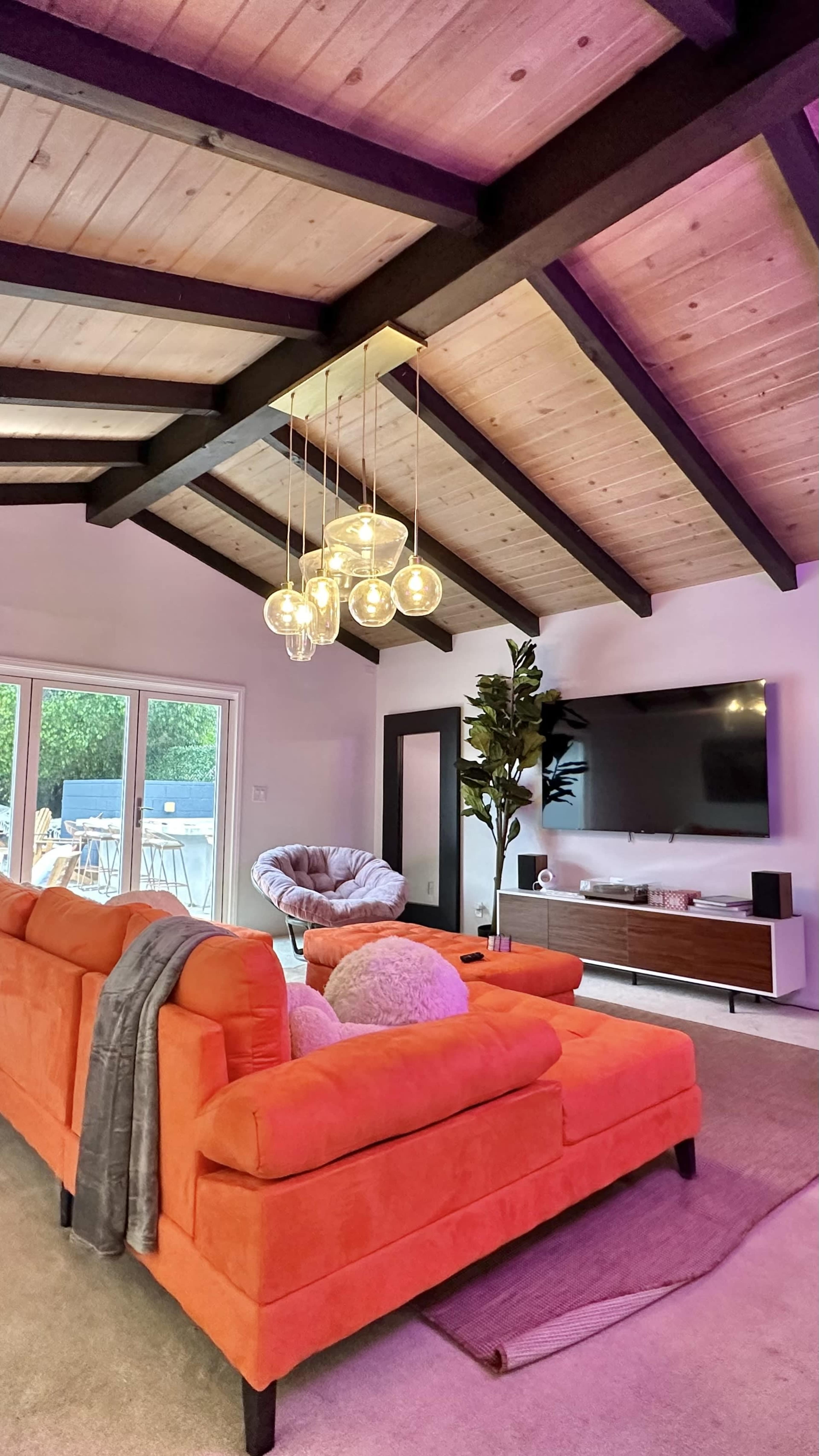 A modern living room with an orange sofa, a TV mounted on the wall, and a chandelier with glass bulbs hanging from the vaulted wooden ceiling.