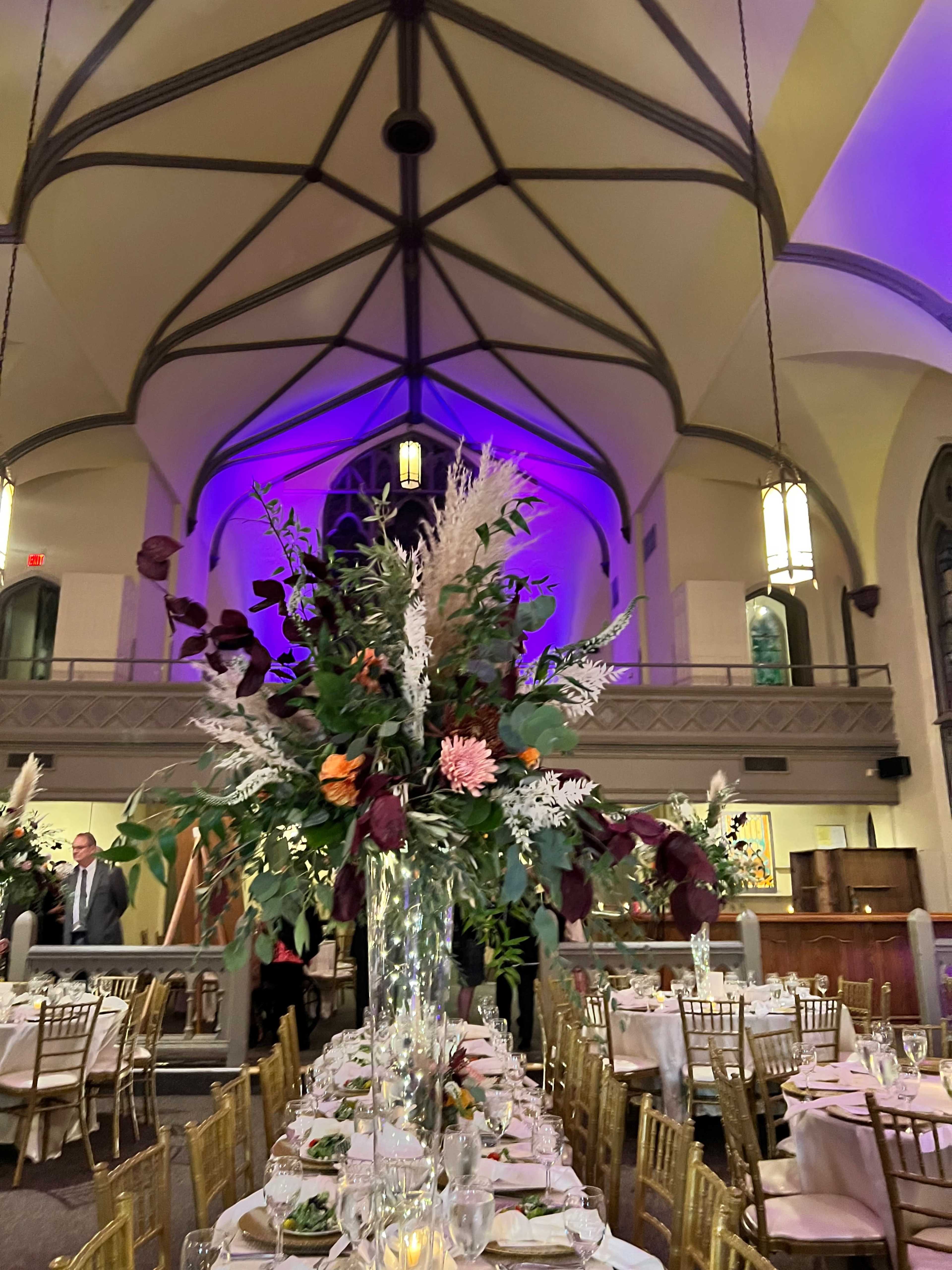 The image shows a dining setup in a hall with tall floral centerpieces and an illuminated ceiling.