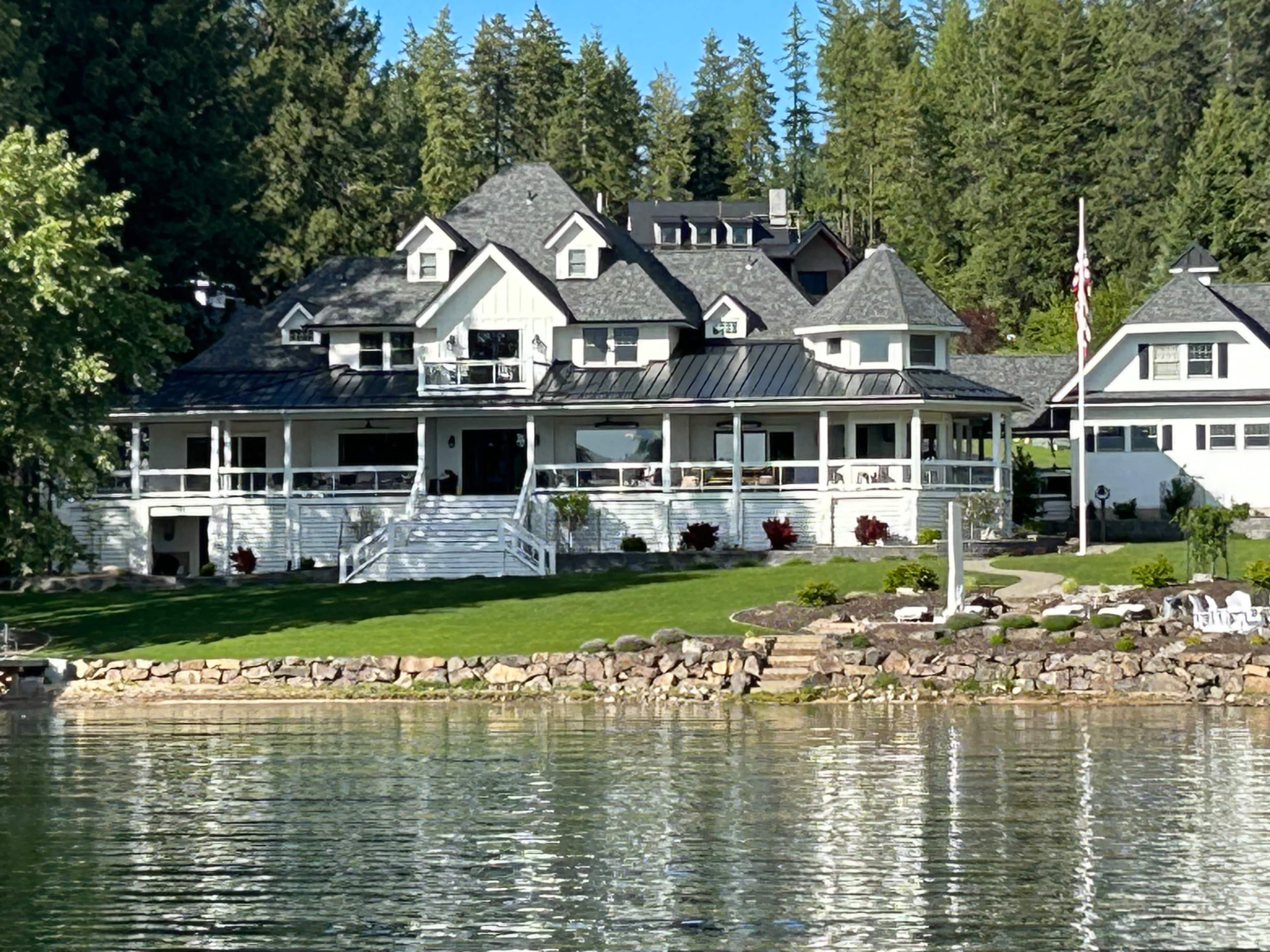 A large, multi-story house with a gray shingled roof and white facade is situated near a lake, surrounded by green trees and a well-maintained lawn.