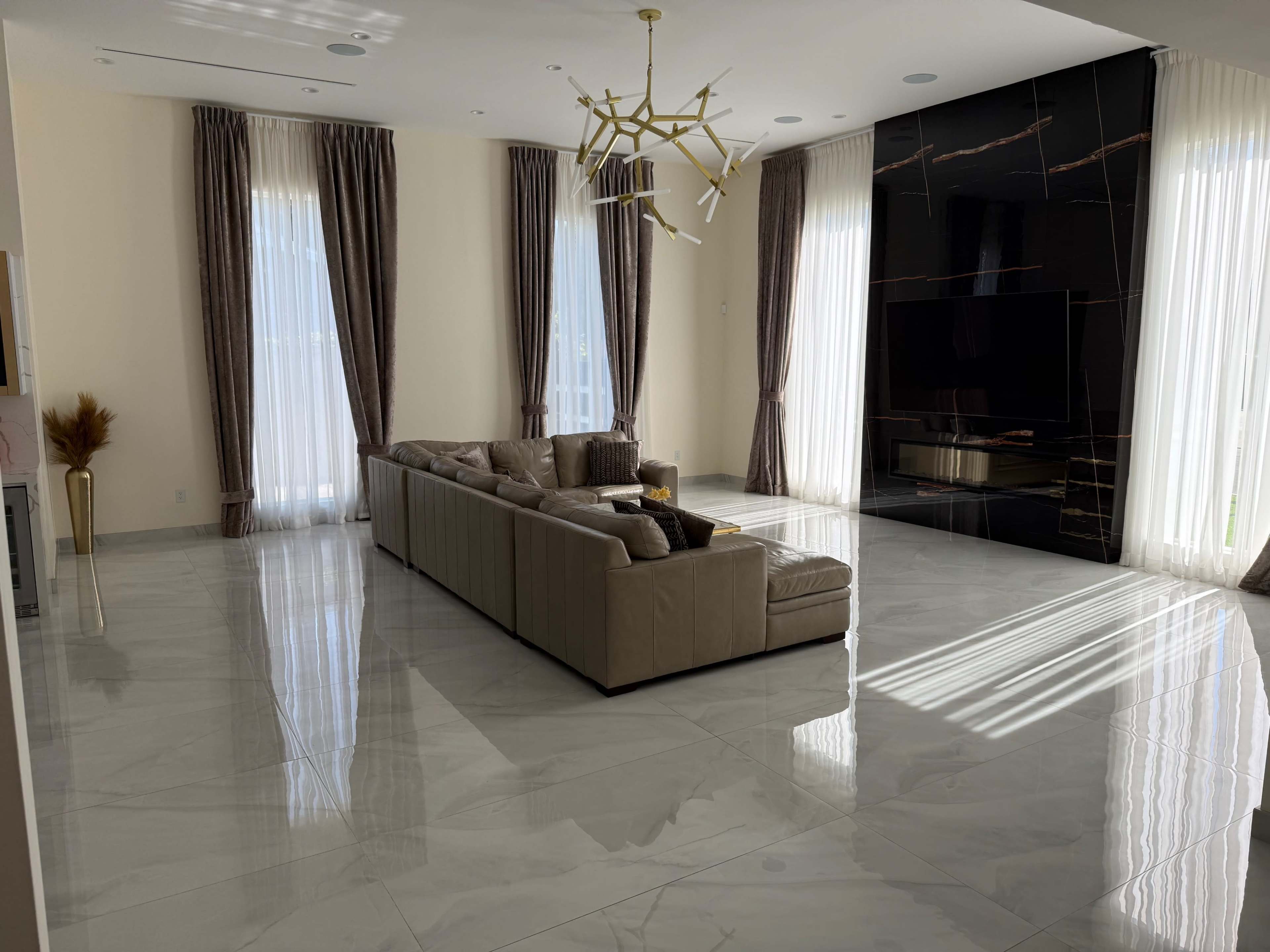The image shows a spacious living room with a large sectional sofa, tall windows dressed in curtains, and a black accent wall featuring a fireplace.