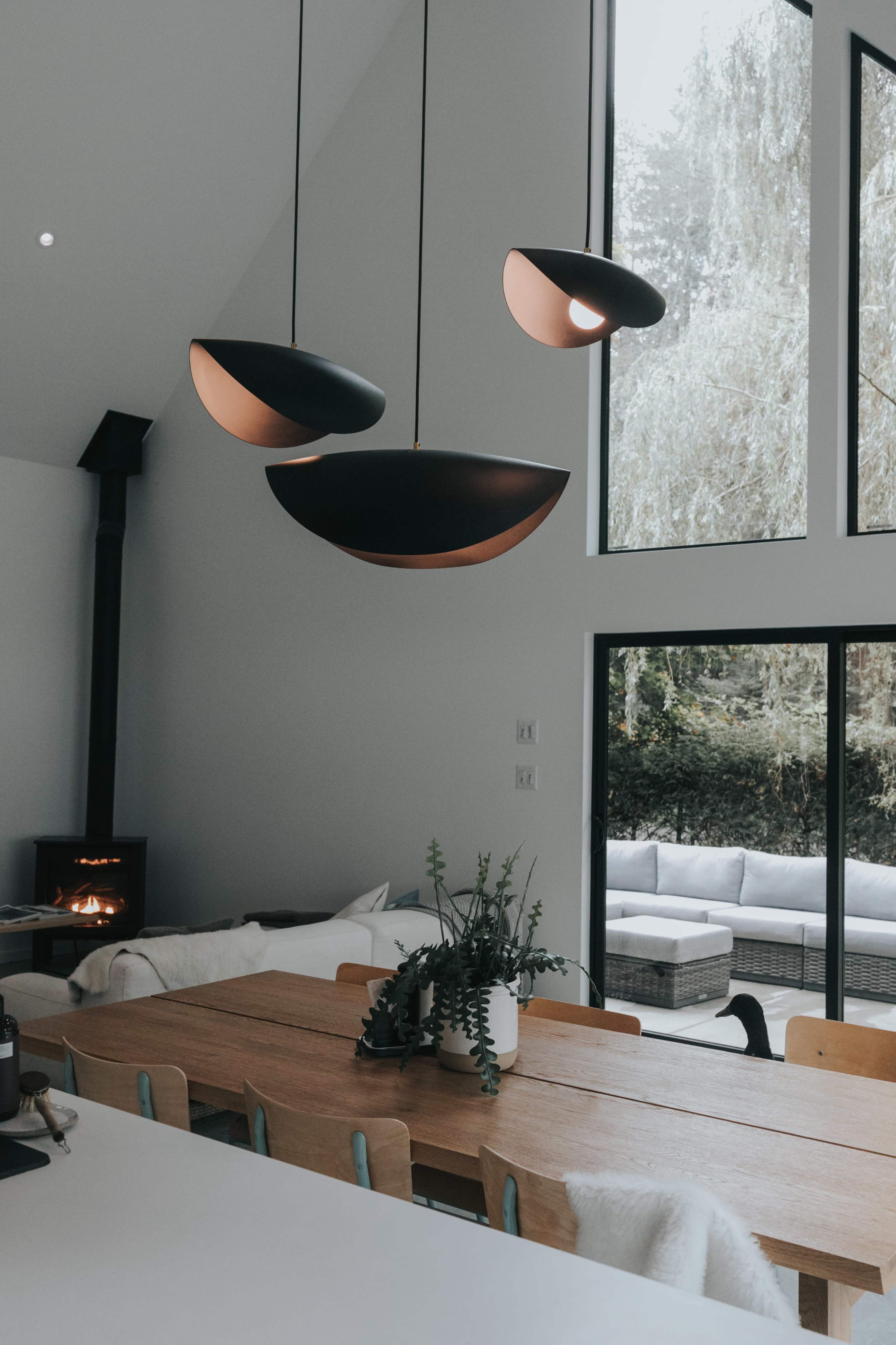 A modern dining area with a long wooden table, three black pendant lights hanging above, and a large window overlooking a patio with seating.