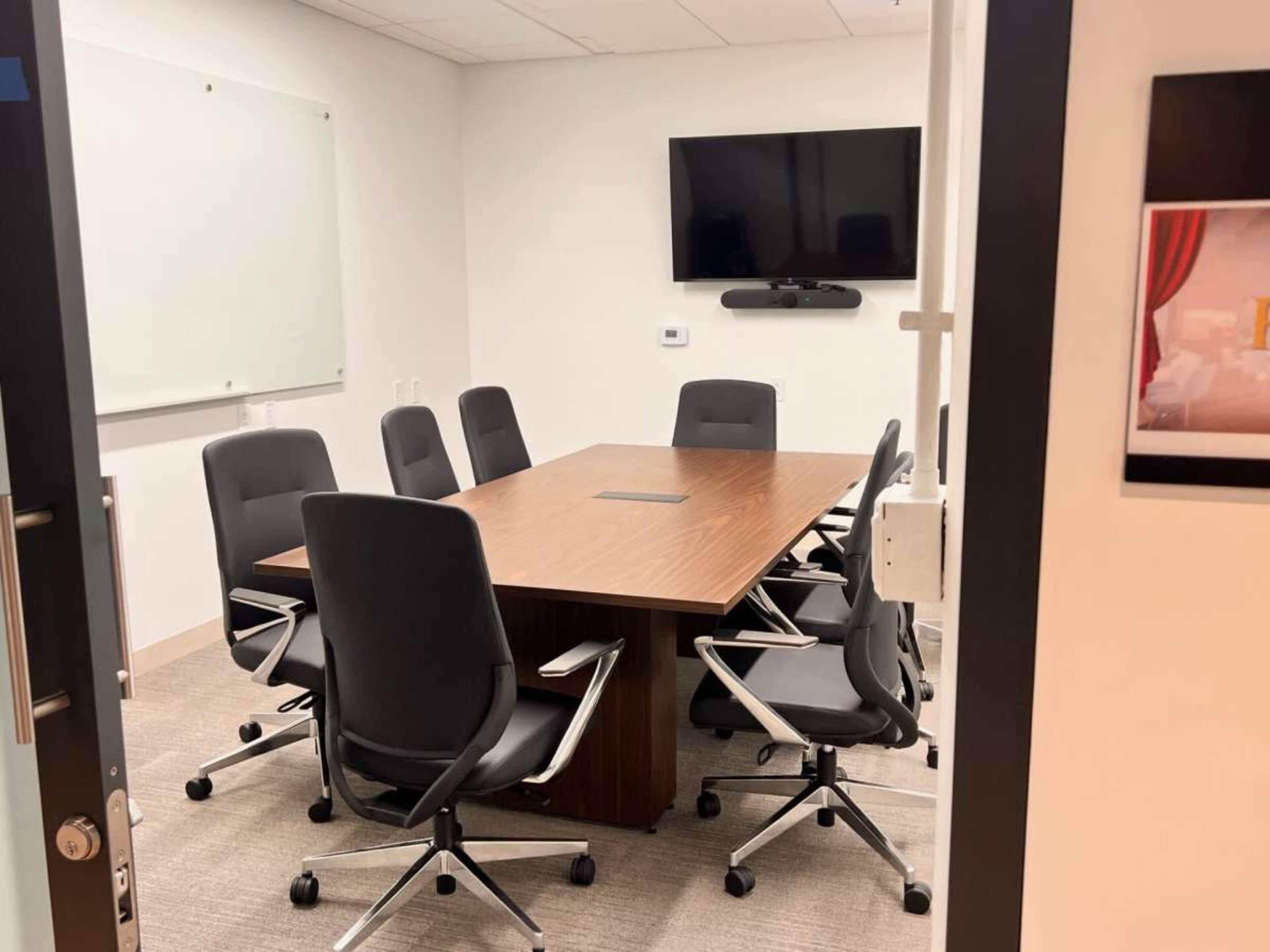 The image shows a conference room with a large table surrounded by six ergonomic chairs and a wall-mounted television.