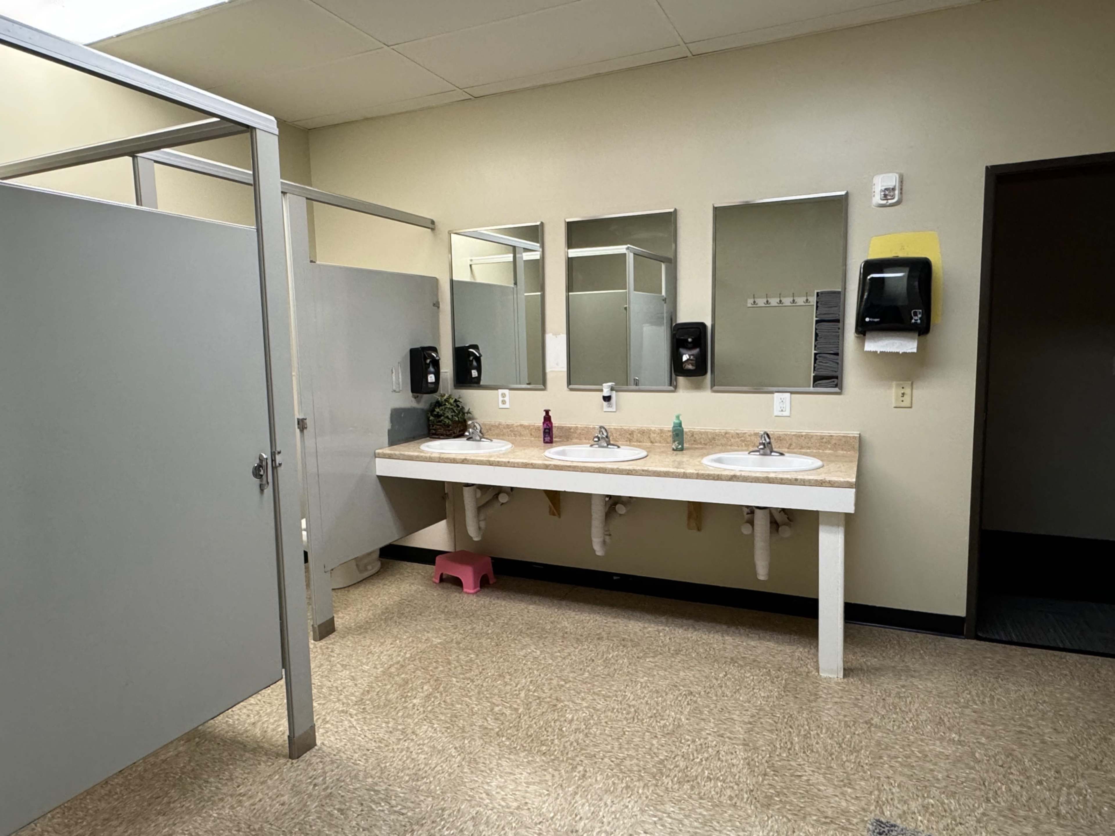 A clean public restroom with two sinks, three mirrors, and two stalls separated by partitions.