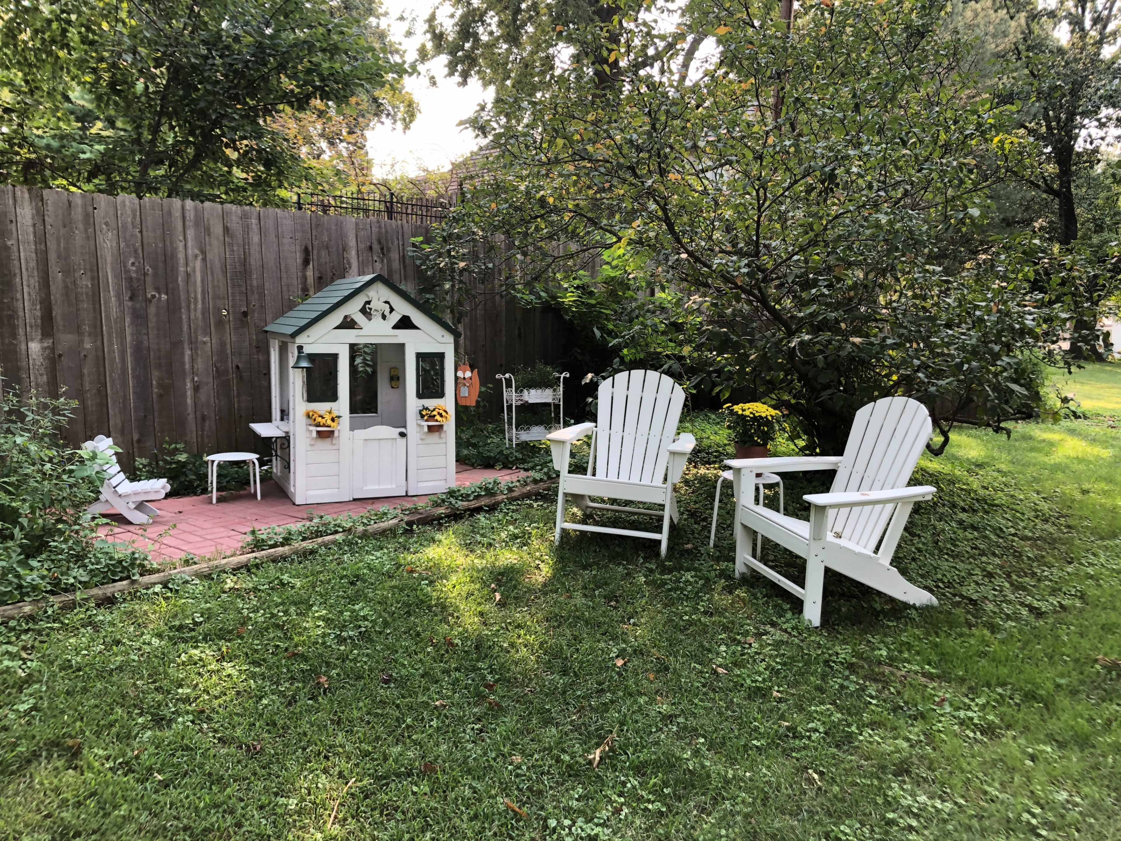 The image shows two white Adirondack chairs facing a small white playhouse in a grassy backyard surrounded by a wooden fence and trees.