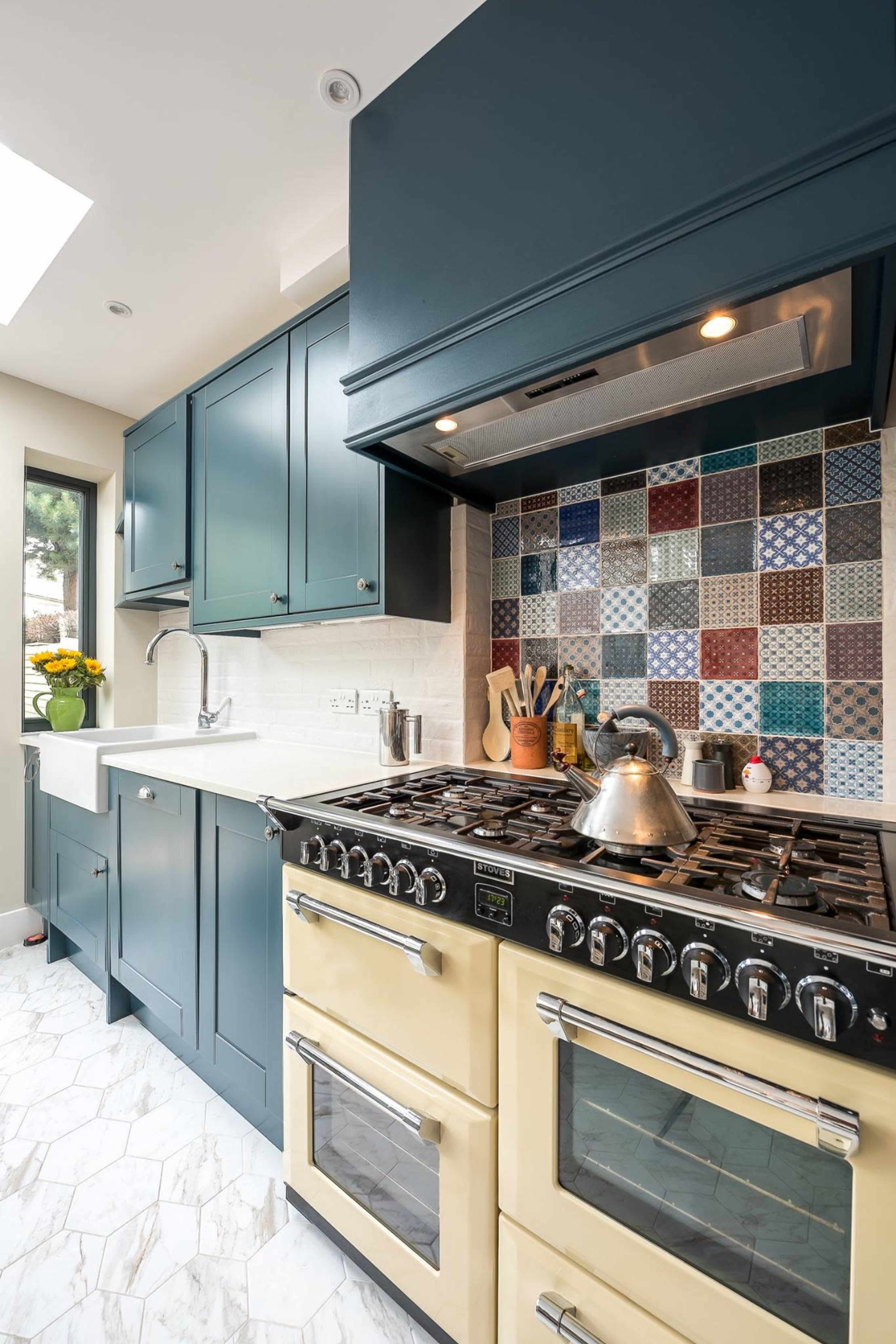 A kitchen features dark blue cabinetry, a tiled backsplash with various patterns, and a yellow gas range with a stainless steel hood.