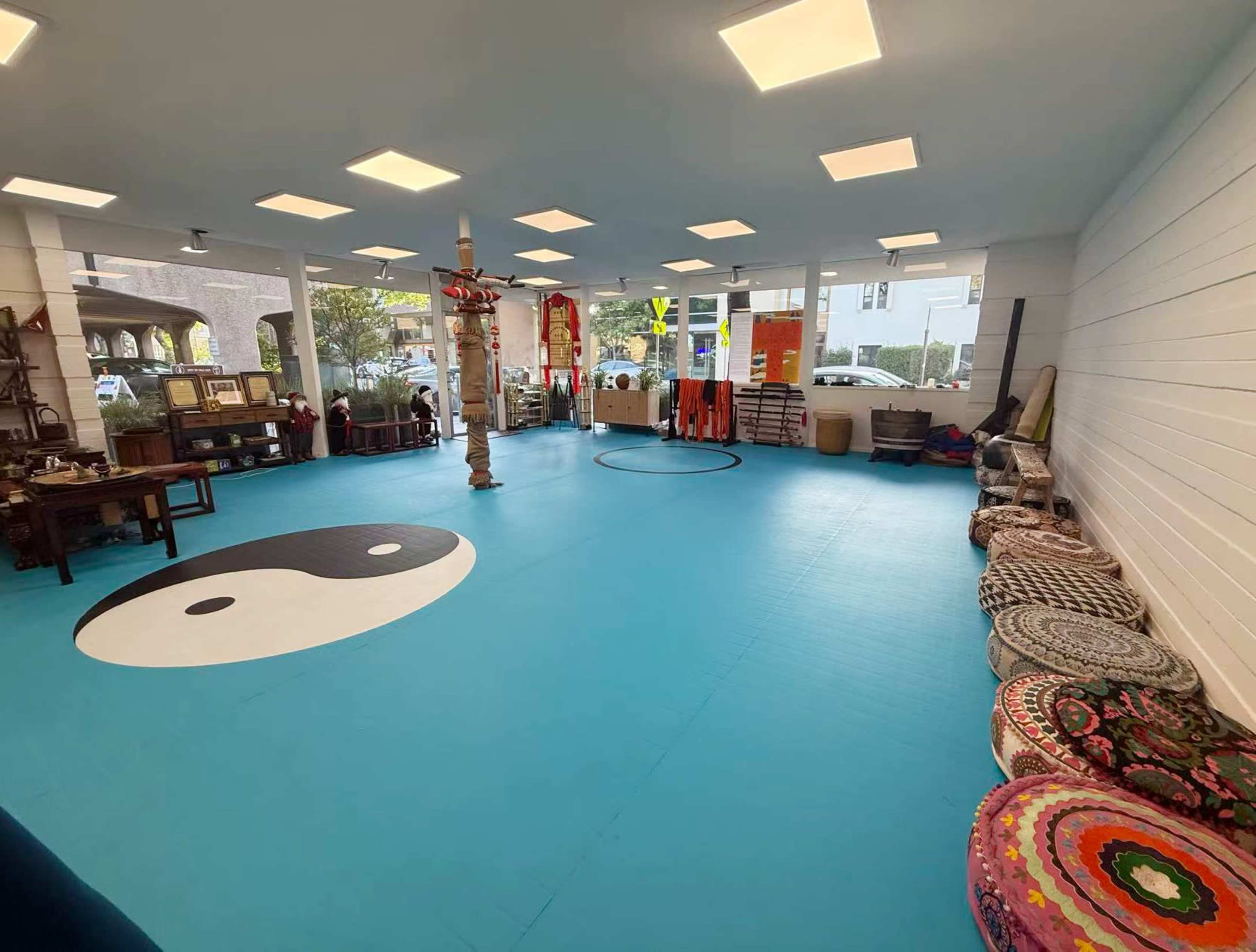 The image shows a spacious room with a blue rubber floor featuring a yin-yang symbol, surrounded by various seating cushions and furniture, with large windows allowing natural light to enter.
