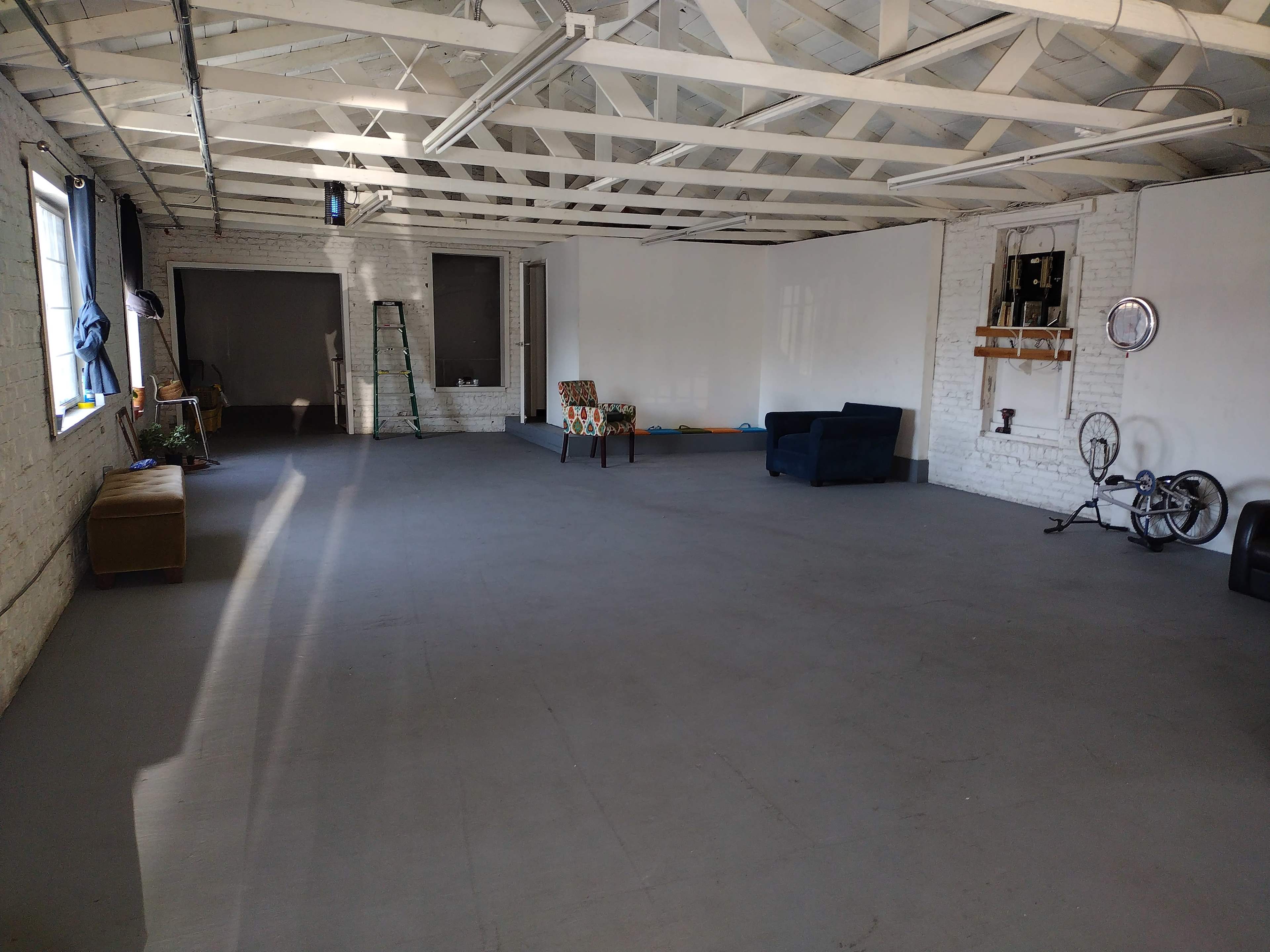 A spacious, empty room with a white brick wall, wooden beams, and minimal furniture including a blue couch and a patterned chair.