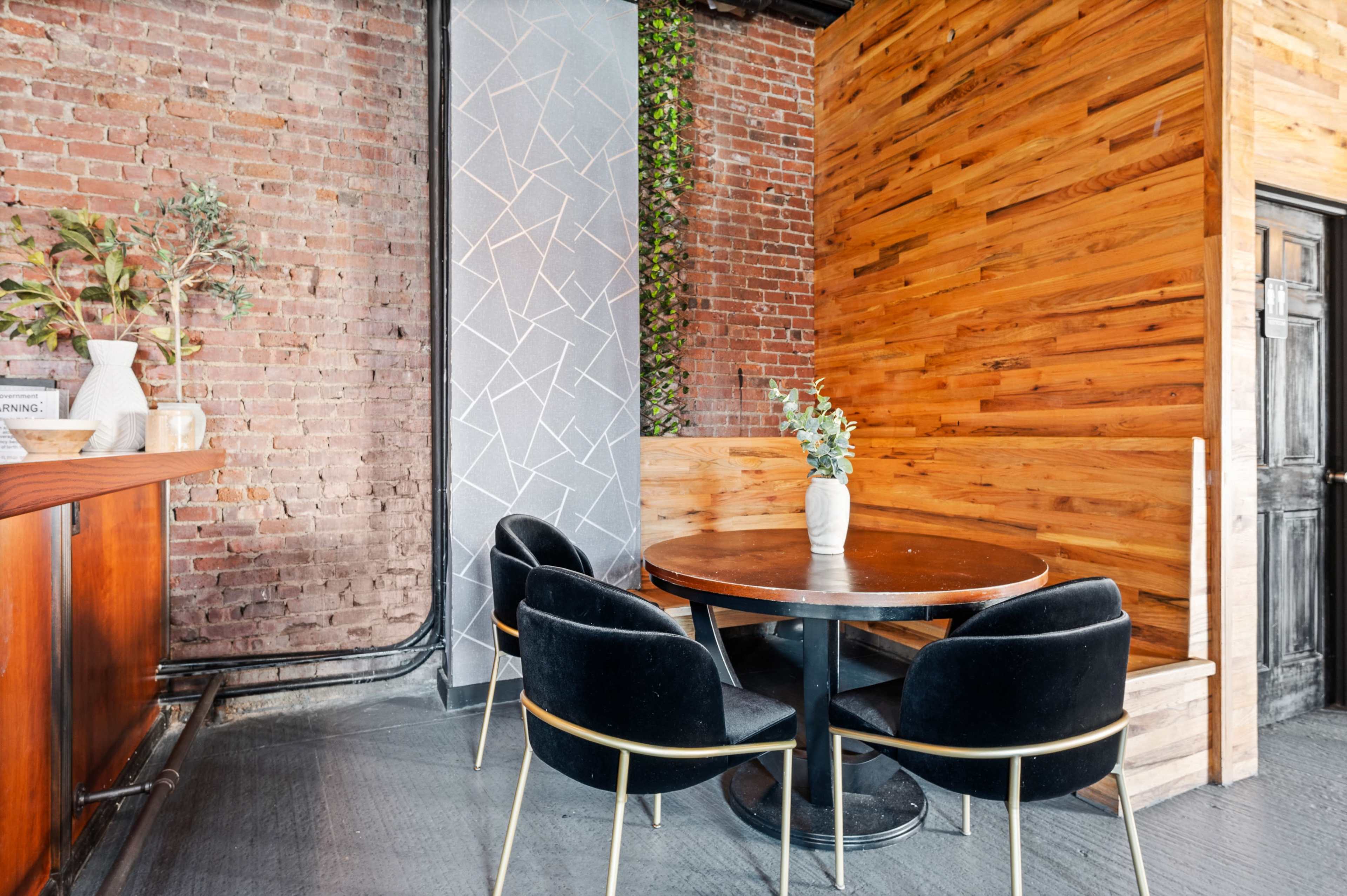 A small dining area features a round black table with four black chairs against a backdrop of exposed brick and wooden walls.