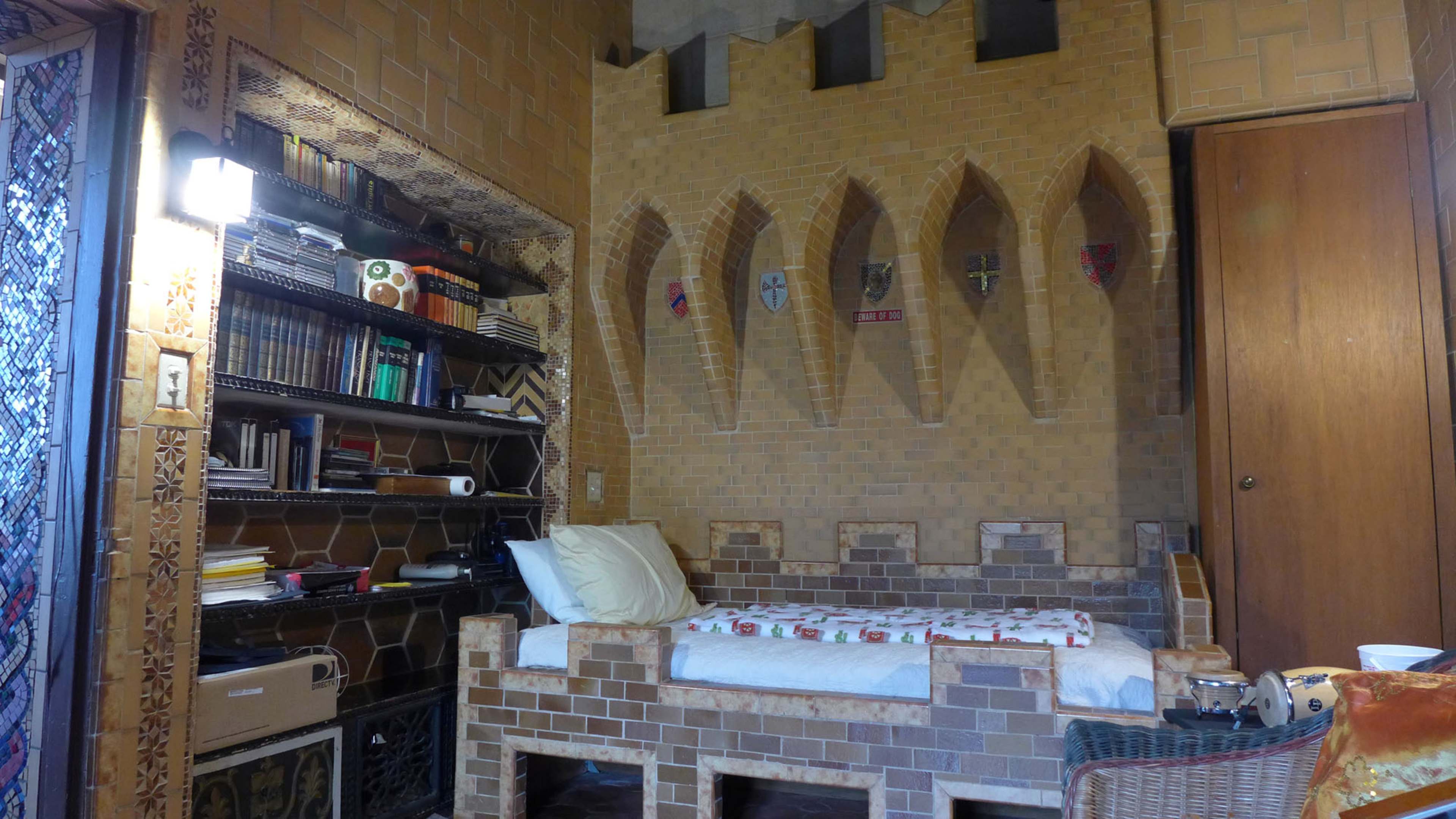The image depicts a small room with a bed made of brick, a wall featuring decorative niches, and shelves filled with books.
