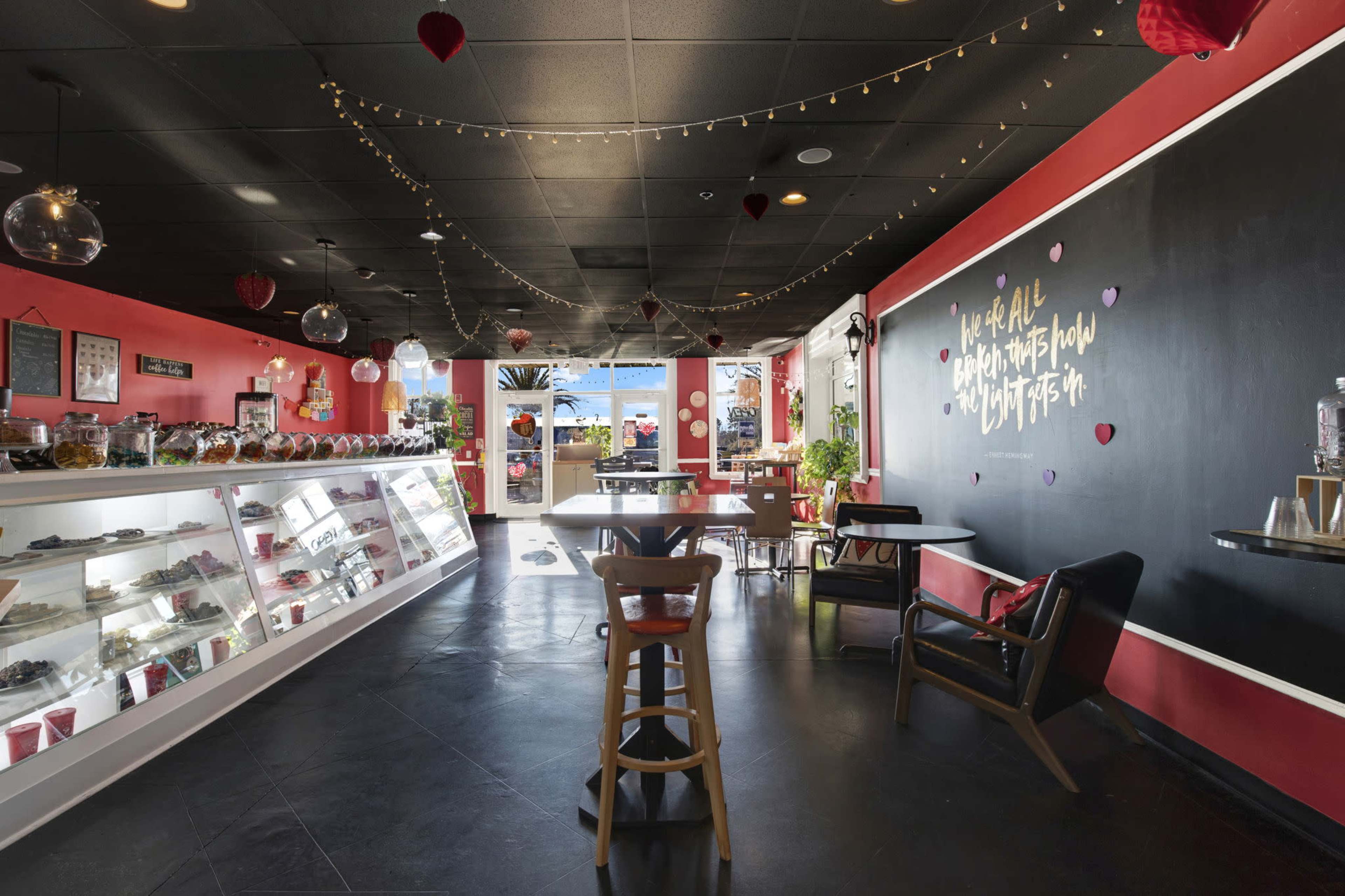 The image shows a cozy café interior with red walls, a display case of treats, string lights, and decorative hearts hanging from the ceiling.
