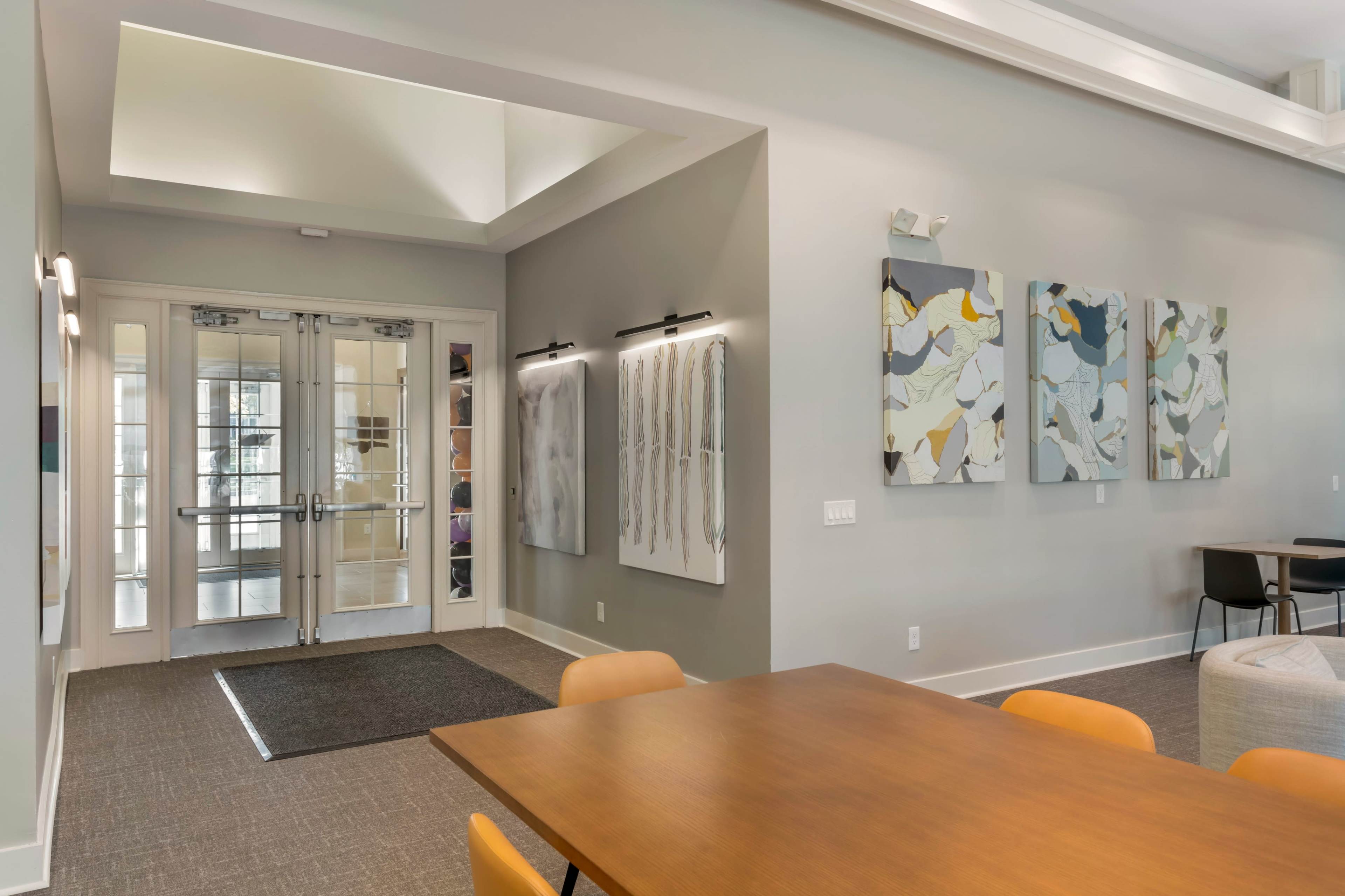 The image shows a spacious indoor area featuring two sets of glass doors, abstract artwork on the walls, and a wooden table with orange chairs.