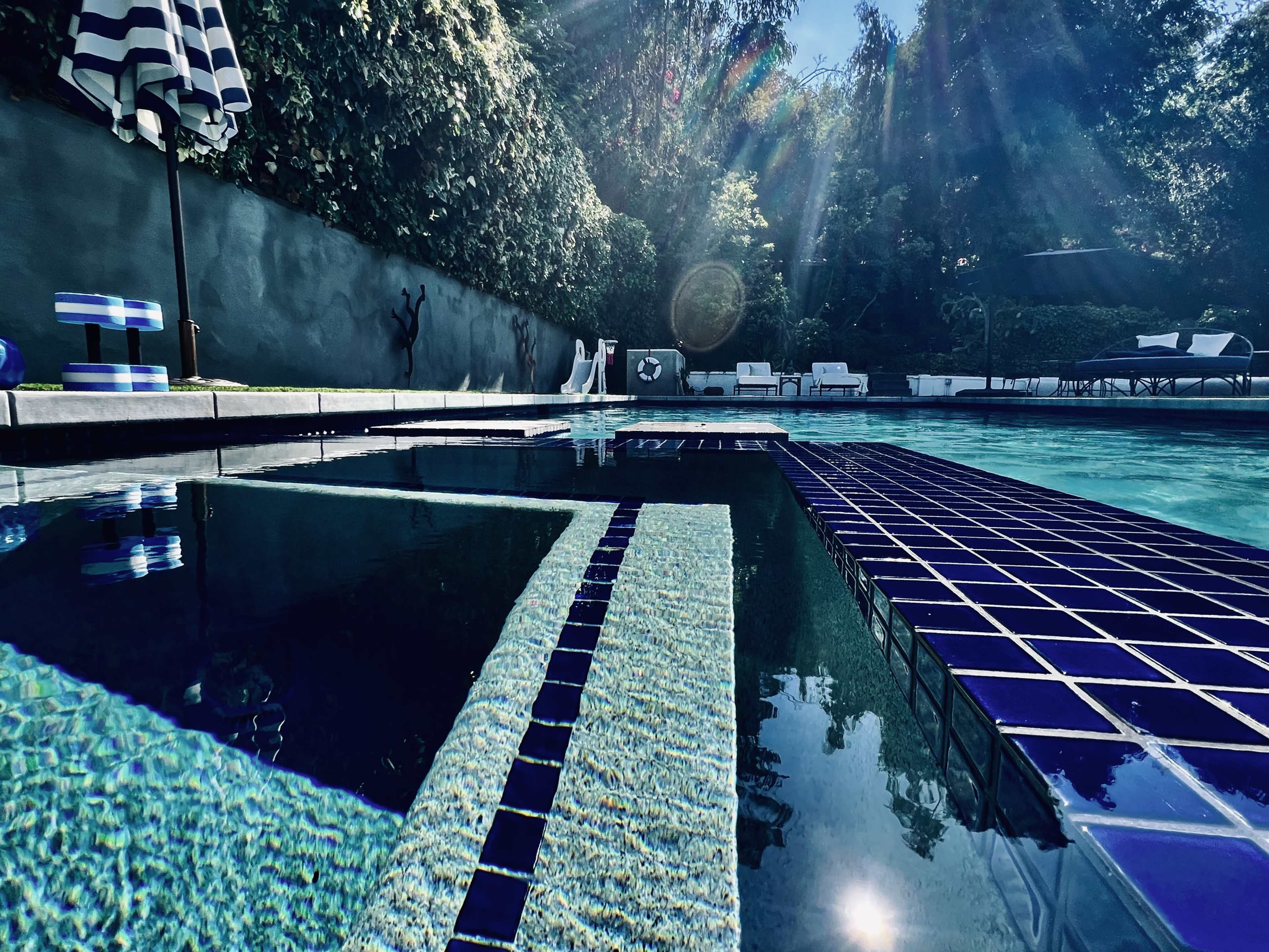 A swimming pool with blue tiles, surrounded by foliage and lounge chairs under a bright sky.