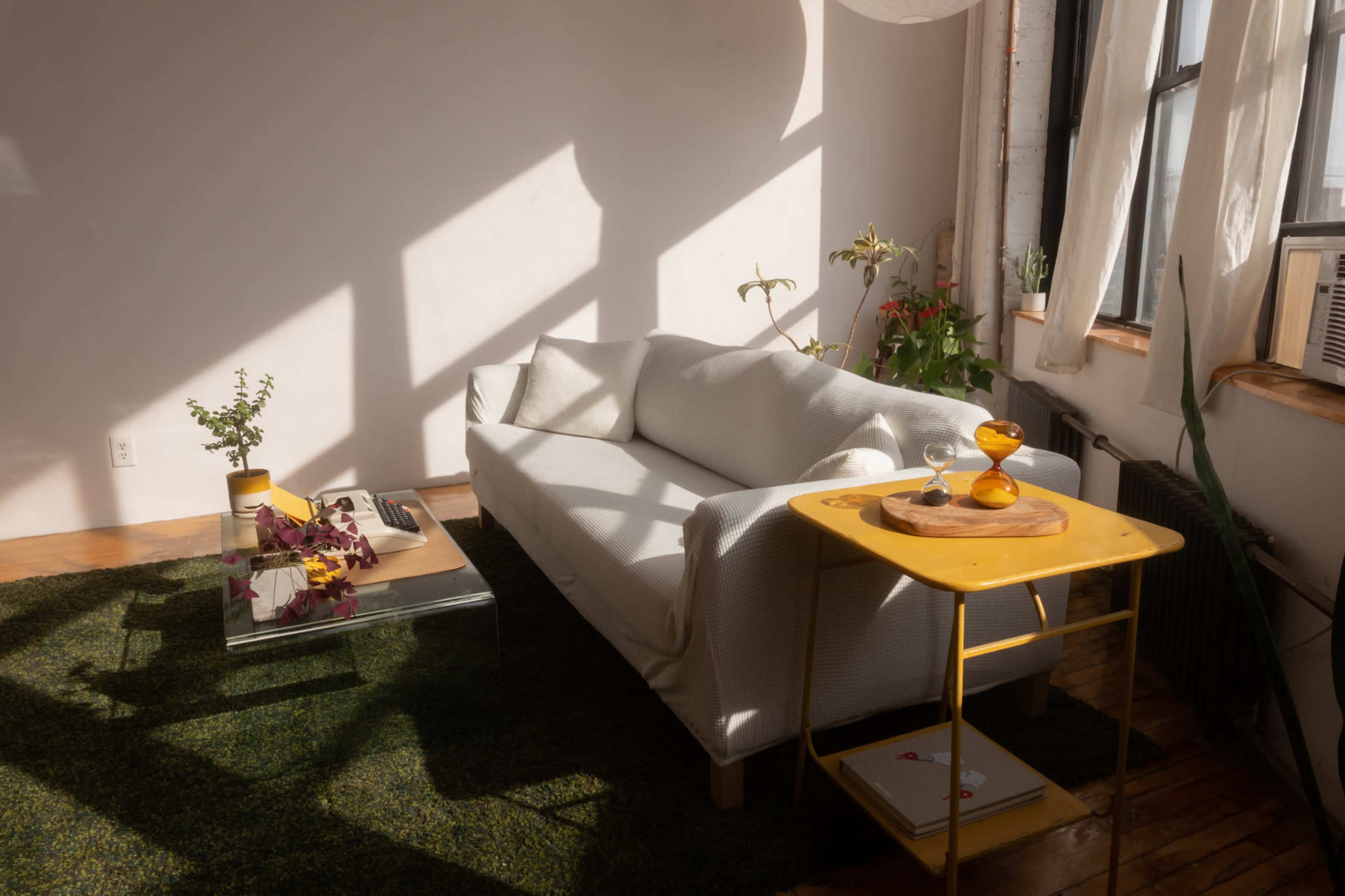 A white sofa is positioned in a sunlit room with plants and a yellow side table holding refreshments.