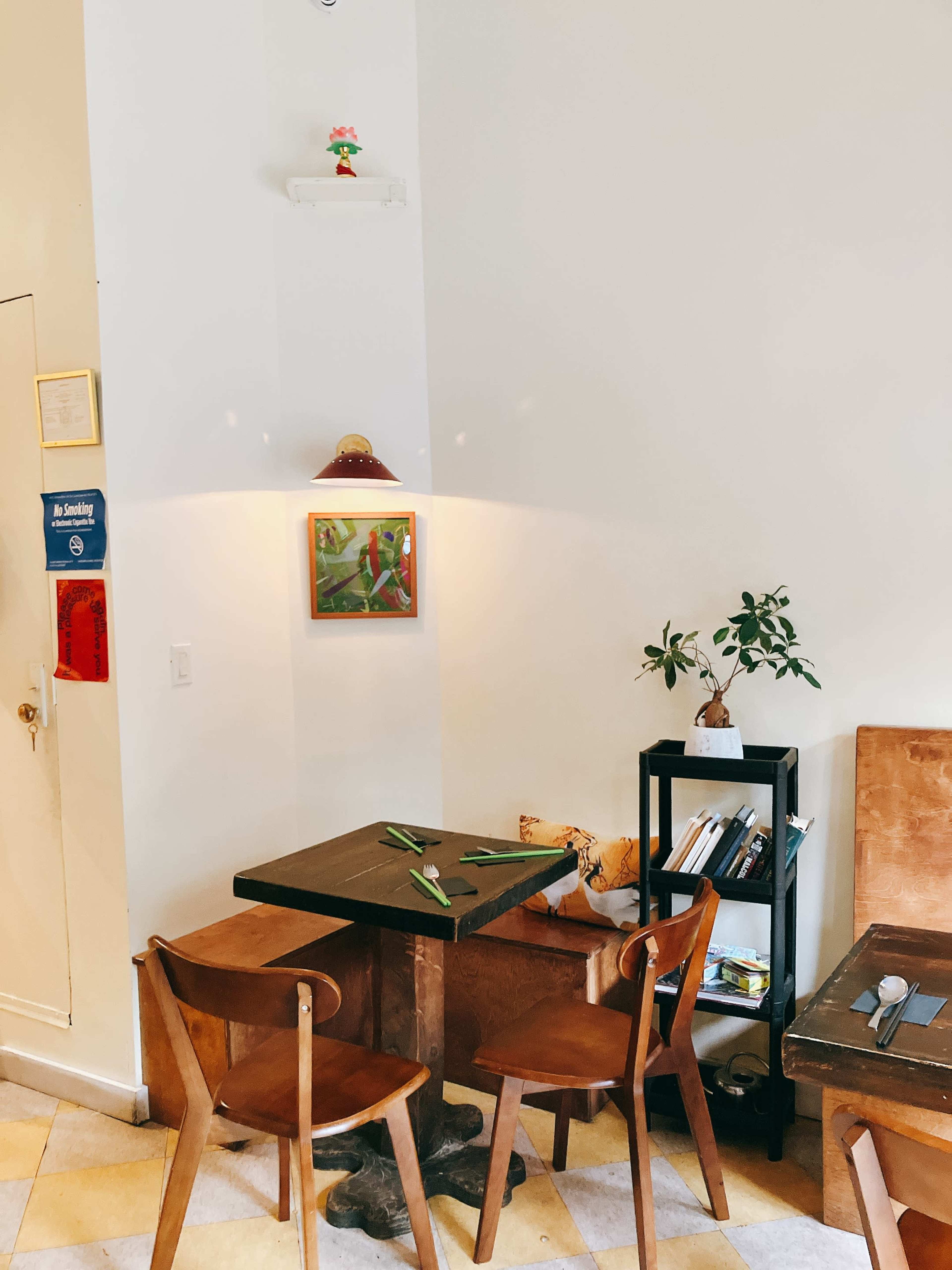 A small café corner features a dark table with two wooden chairs, a potted plant, and a colorful painting hanging on the wall above.