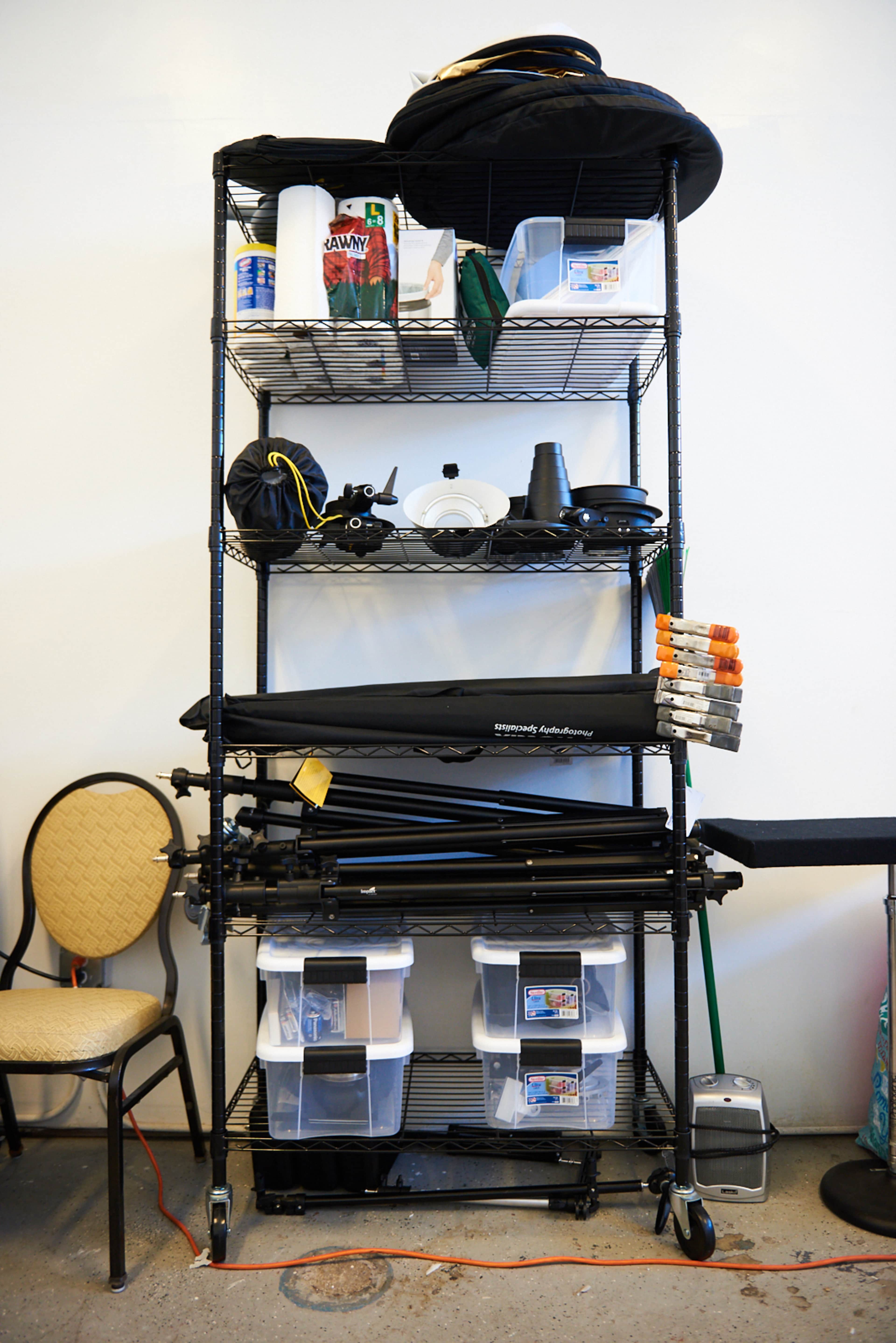 A metal shelving unit is filled with various photography equipment, including light stands, black backdrops, and storage bins, next to a chair.