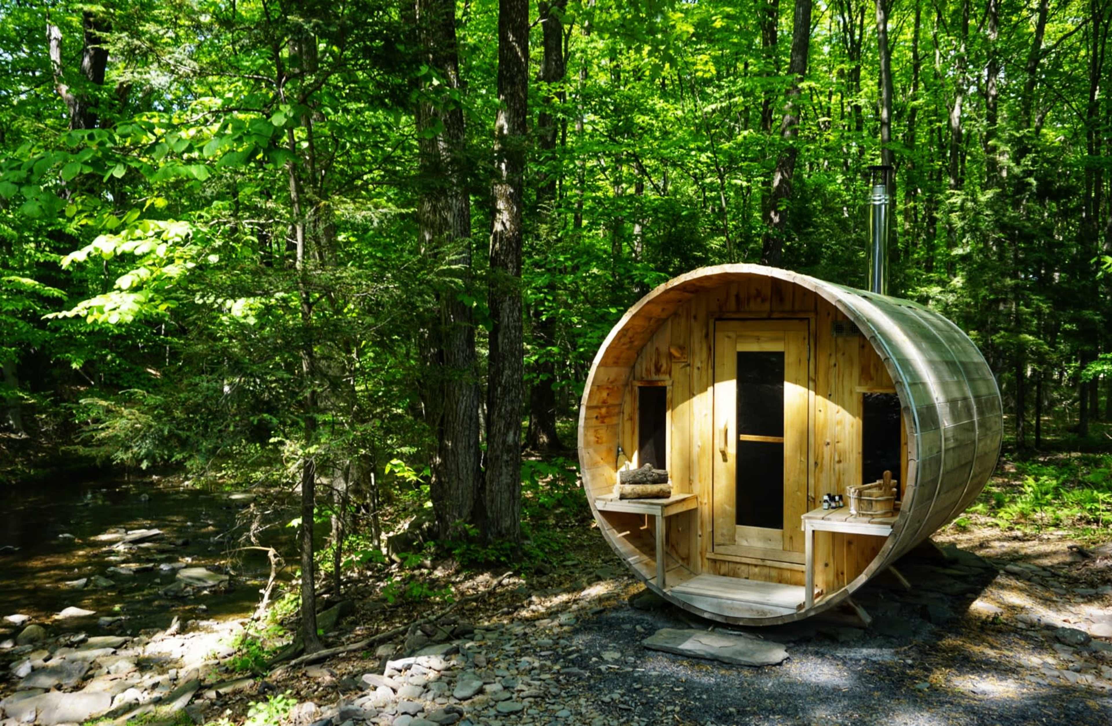 A wooden, cylindrical cabin is located in a forest by a small stream, surrounded by lush green trees.