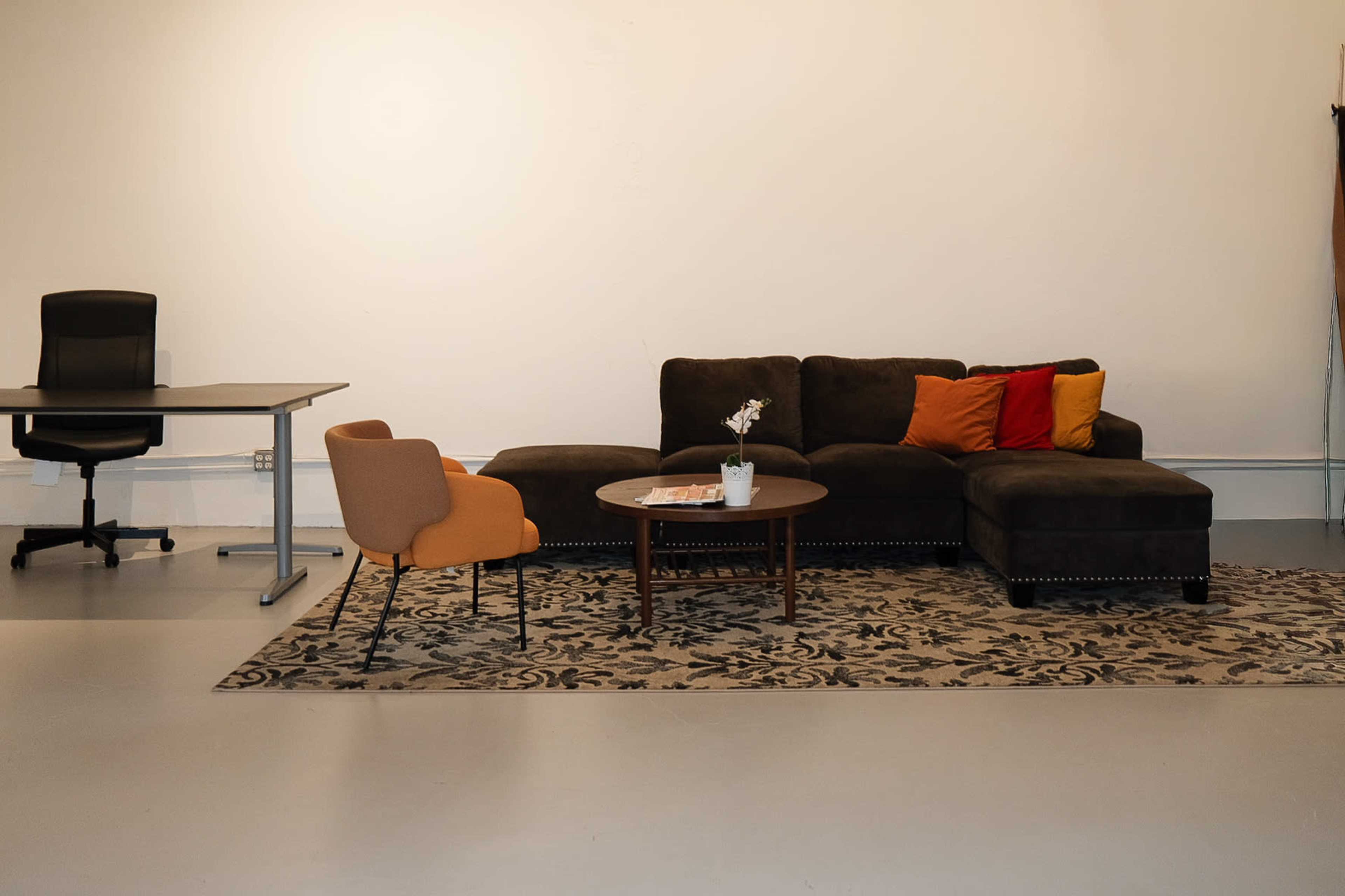 The image depicts a modern living room setup featuring a dark sectional sofa with colorful cushions, a round coffee table with a plant, an orange accent chair, and a desk with a rolling office chair.