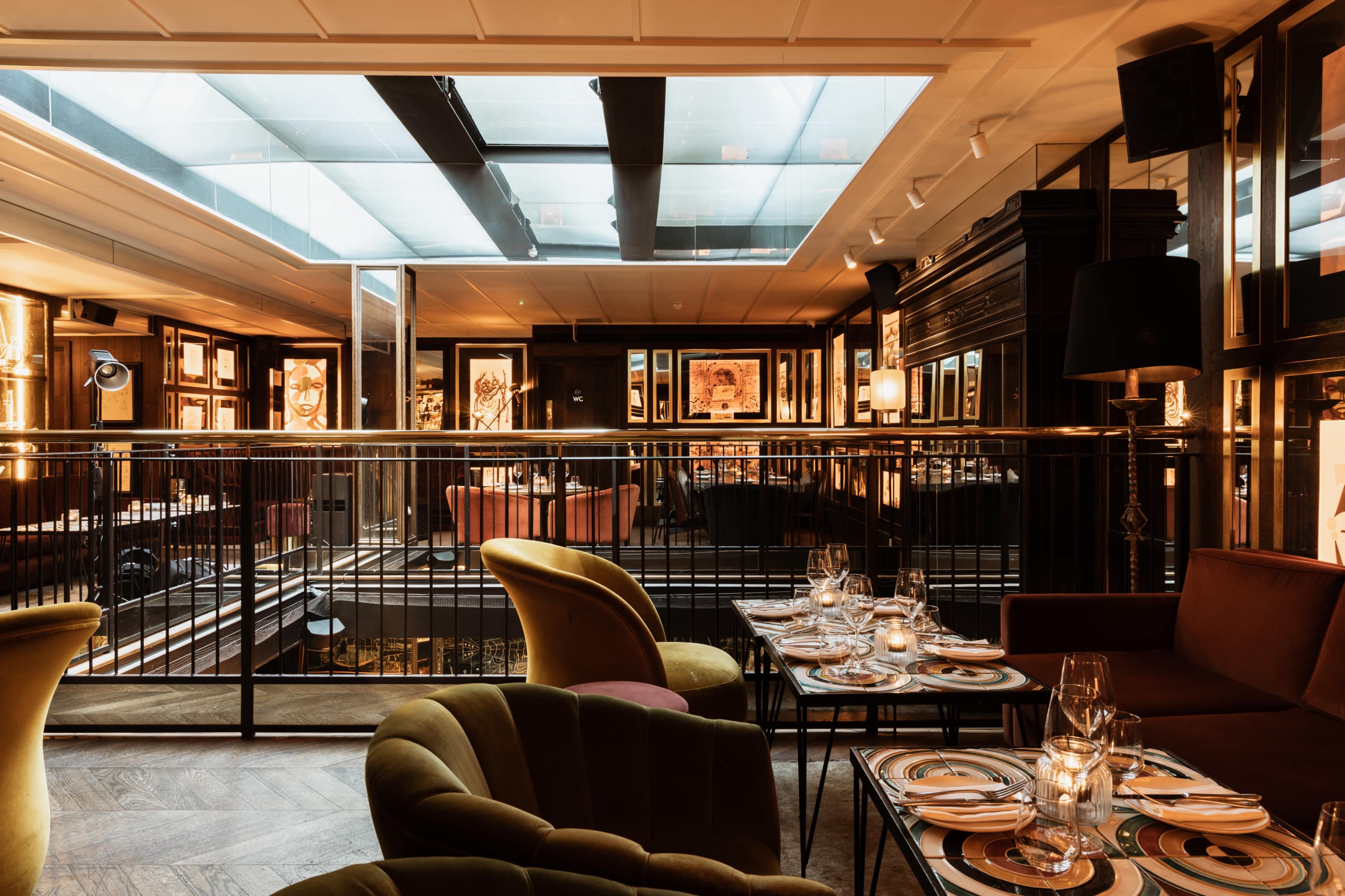A restaurant interior with elegant seating, decorative lighting, and a glass ceiling allowing natural light to filter in.