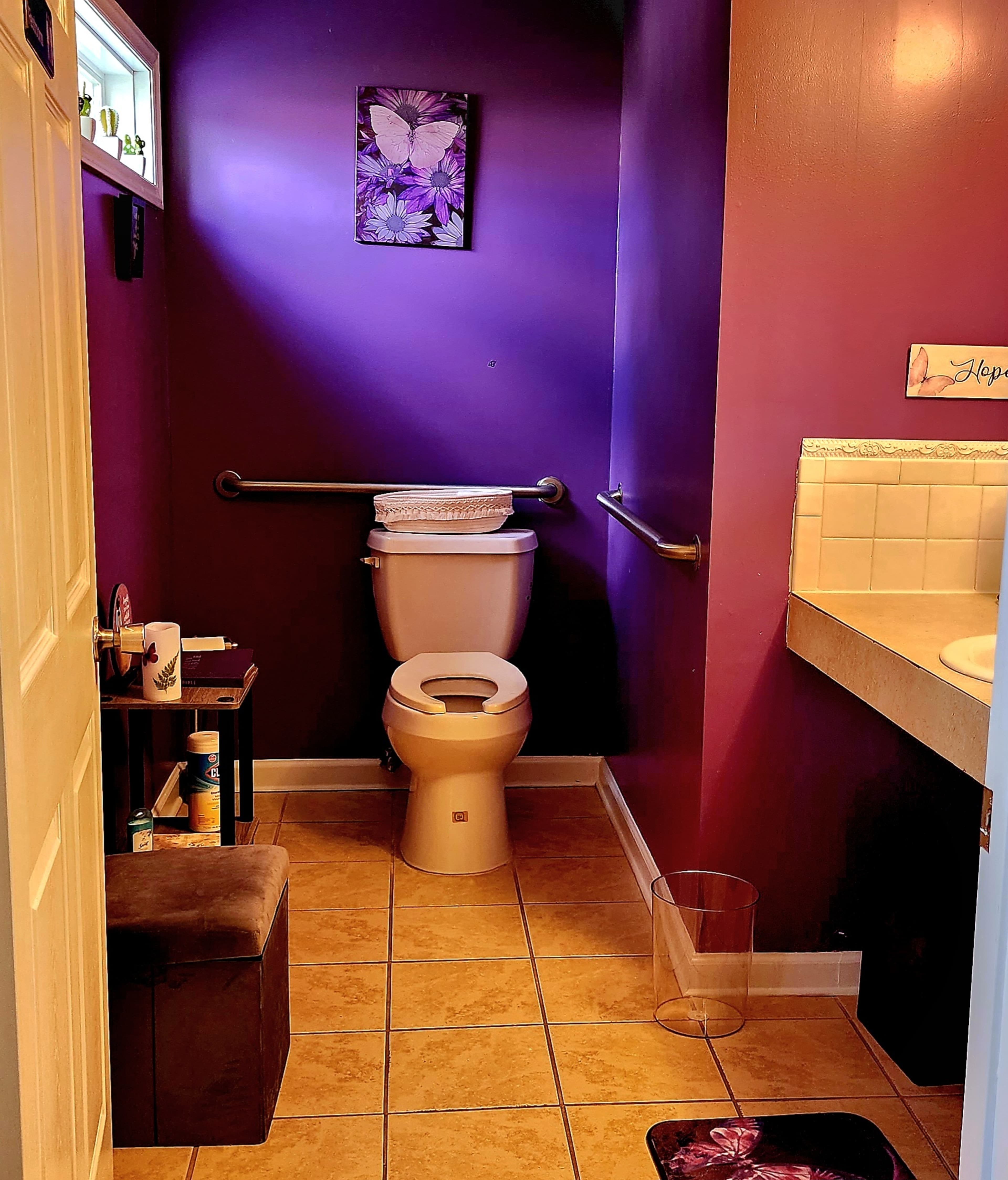 A small bathroom with purple walls, featuring a toilet, a sink, a wastebasket, and a stool beside the door.