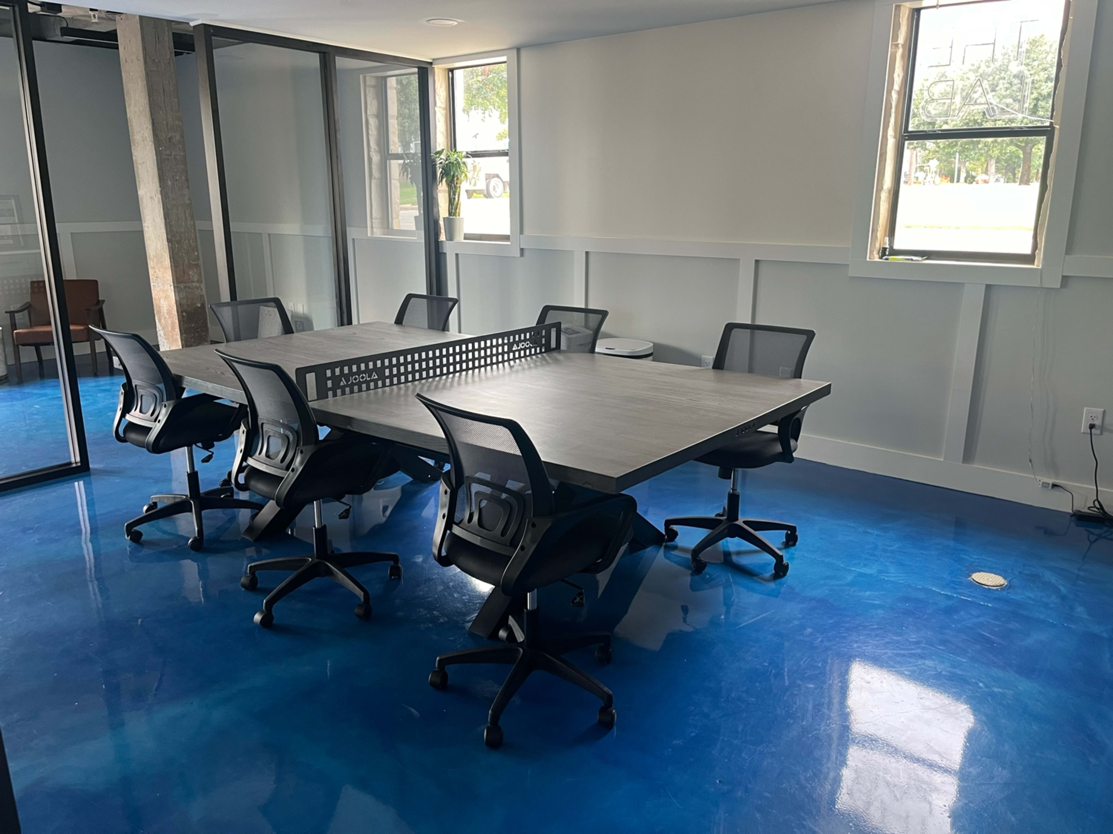 The image shows a conference room with a large table surrounded by six rolling chairs, illuminated by natural light coming through two windows.