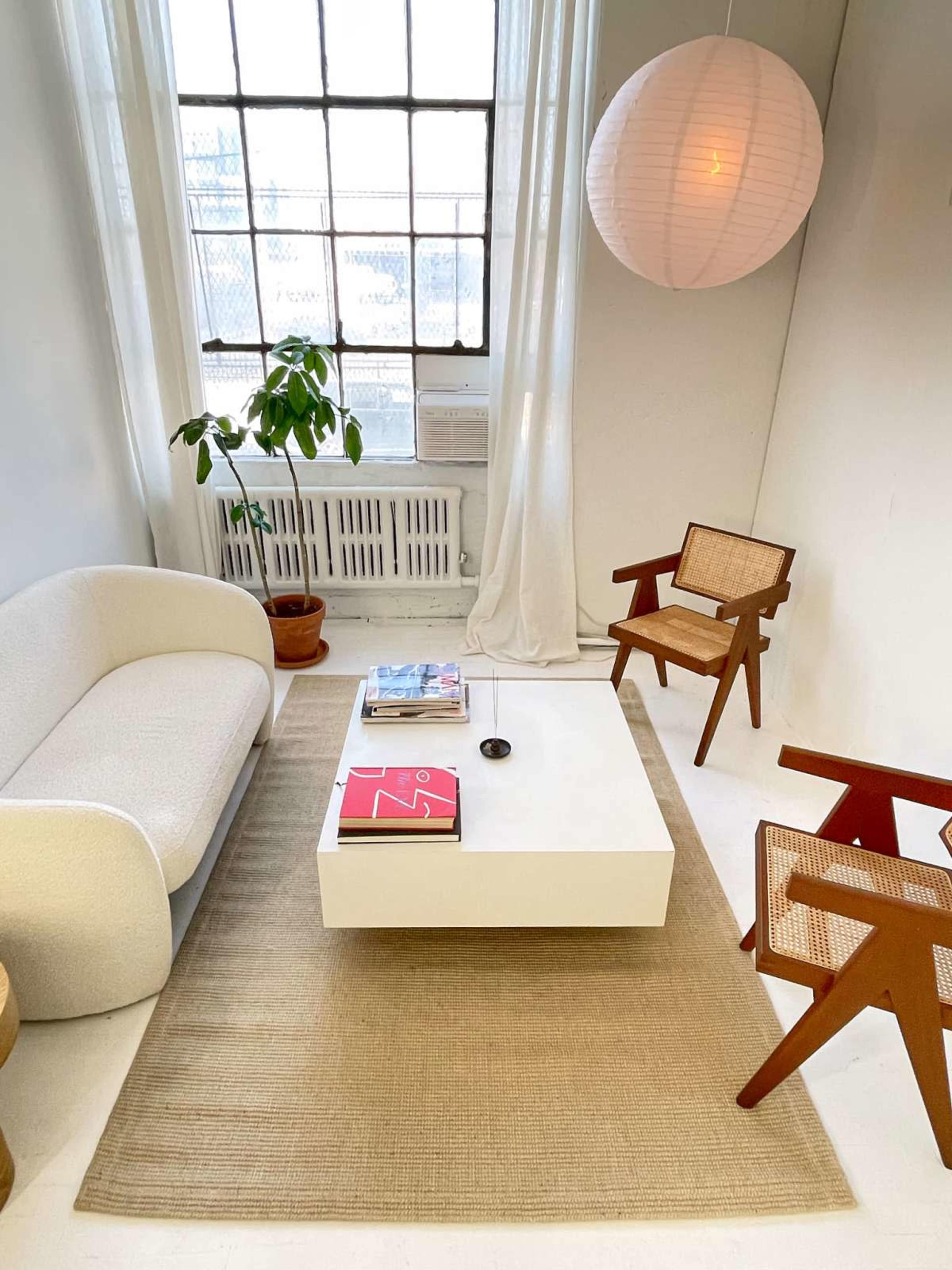 The image shows a minimalist living room with a white couch, a coffee table, two wooden chairs, a plant, and a large window letting in natural light.