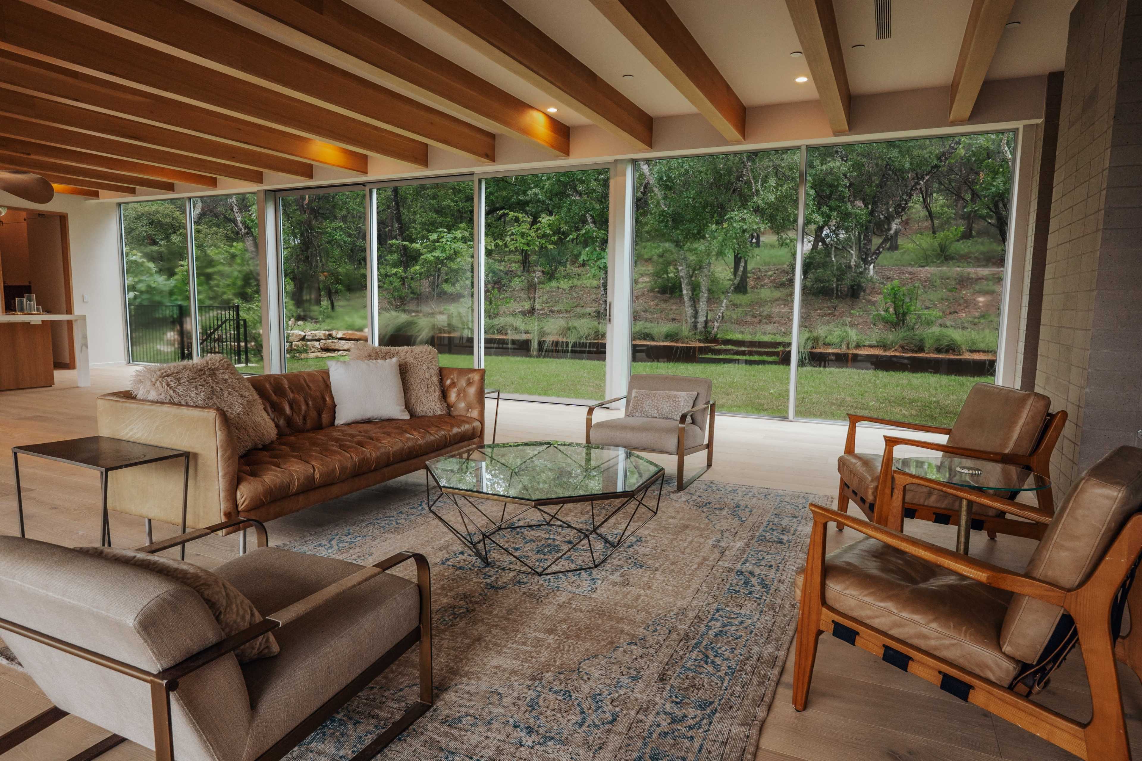 A modern living room features a leather sofa, glass coffee table, and various chairs with large windows providing a view of lush greenery outside.