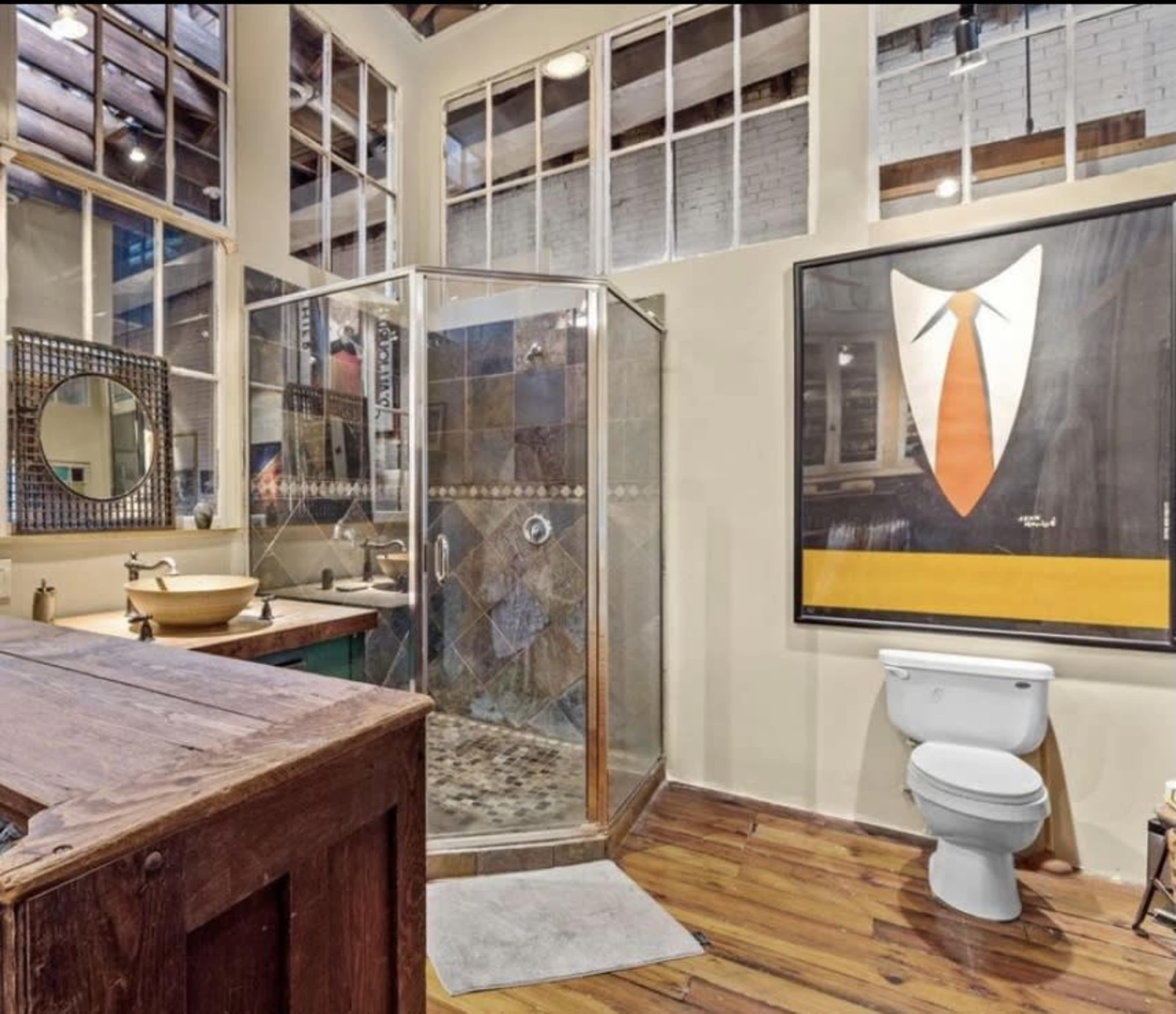 A modern bathroom featuring a glass shower, a vanity with a bowl sink, a toilet, and a large artwork of a suit hanging on the wall.