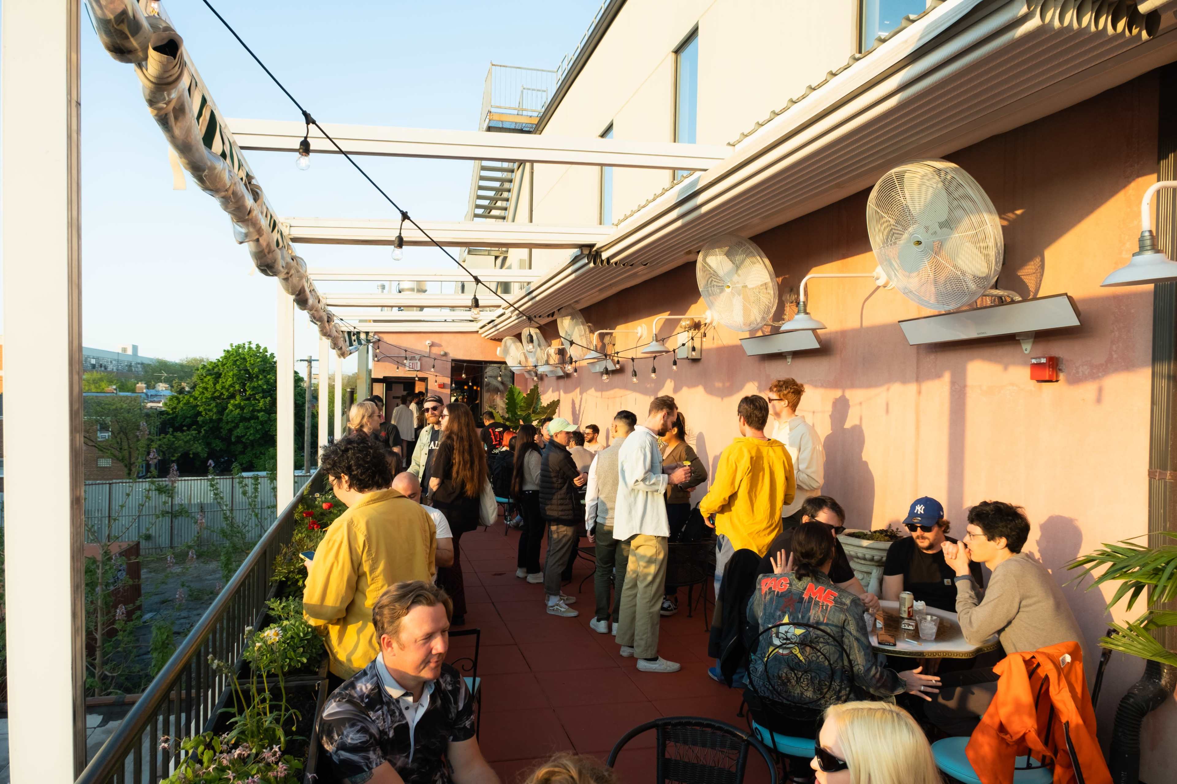 A crowd of people socializes on a rooftop patio with plants and large fans.