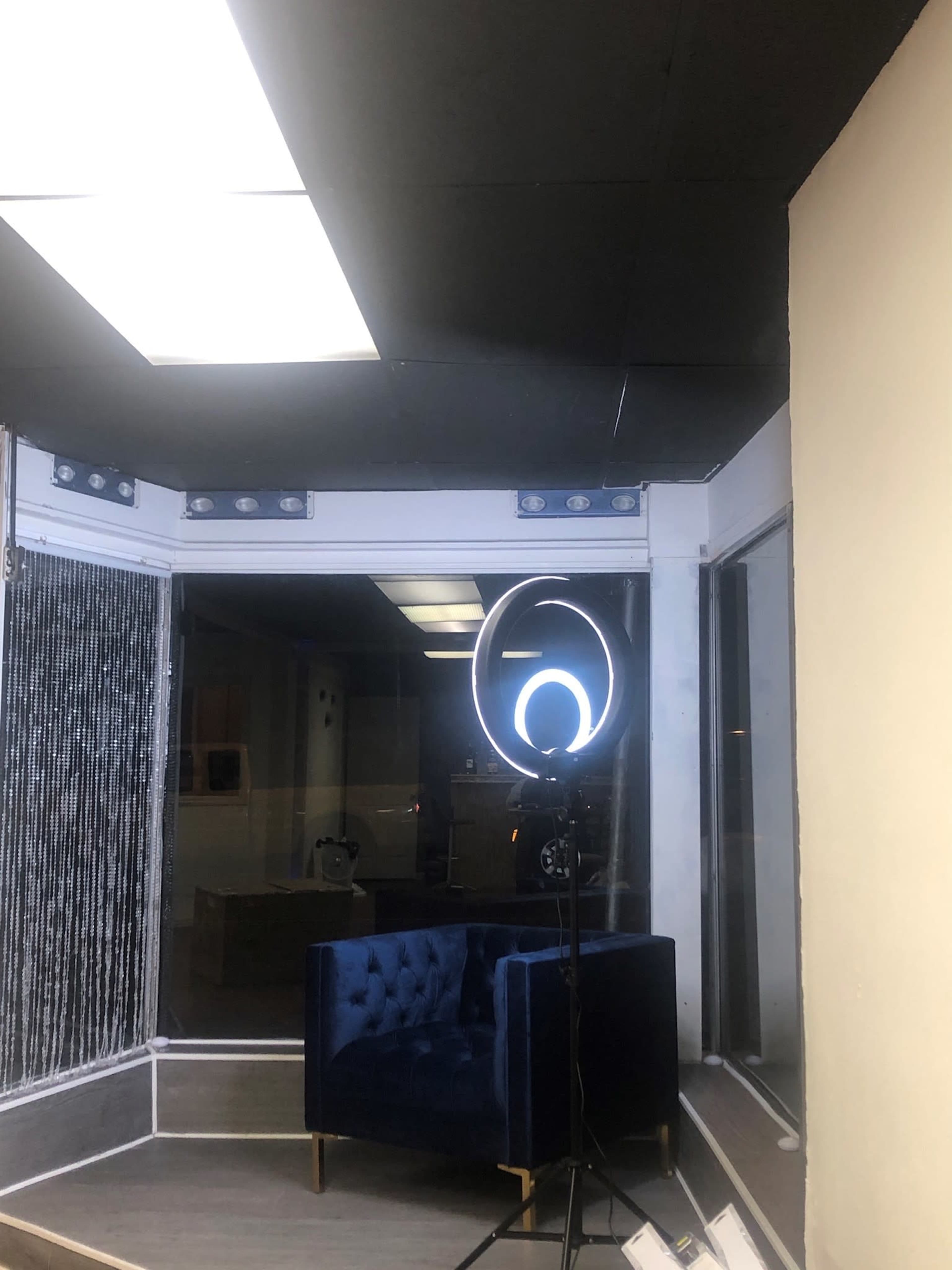 The image shows a corner of a room featuring a blue velvet chair and a ring light on a stand, with large windows displaying a dimly lit outside.